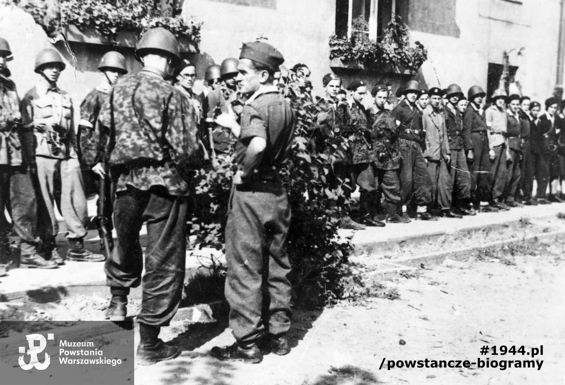 Fotografia z Powstania Warszawskiego. Żoliborz, żołnierze zgrupowania "Żyrafa". Zbiórka plutonu 227 na podwórzu bloku przy ul. Krasińskiego 20. Pierwszy od lewej Ryszard Witrzenc "Wacek", Janusz Noskowski "Zbyszek", w okularach Waldemar Rolbiecki "Popielica"; pierwsza od prawej: Kazimiera Marzec "Kamila", za nią, Sławomir Zawadzki "Lech", Jerzy Szletyński "Gryf", w jasnej bluzie Tymoteusz Duchowski "Motek", Andrzej Tomiak "Kita", Czesław Nowak " Korczak", w jasnej marynarce Jan Kępiński "Ostoja", Bohdan Turos "Milczek", Włodzimierz Krystosiak "Mongoł", Józef Małkowski "Sokół". Przed frontem plutonu stoją: tyłem w panterce plut. pchor. Andrzej Wiczyński "Antek", profilem w furażerce por. Ryszard Wołczyński "Tatar". Fot. ze zbiorów Muzeum Powstania Warszawskiego, sygn. MPW-IH/4264