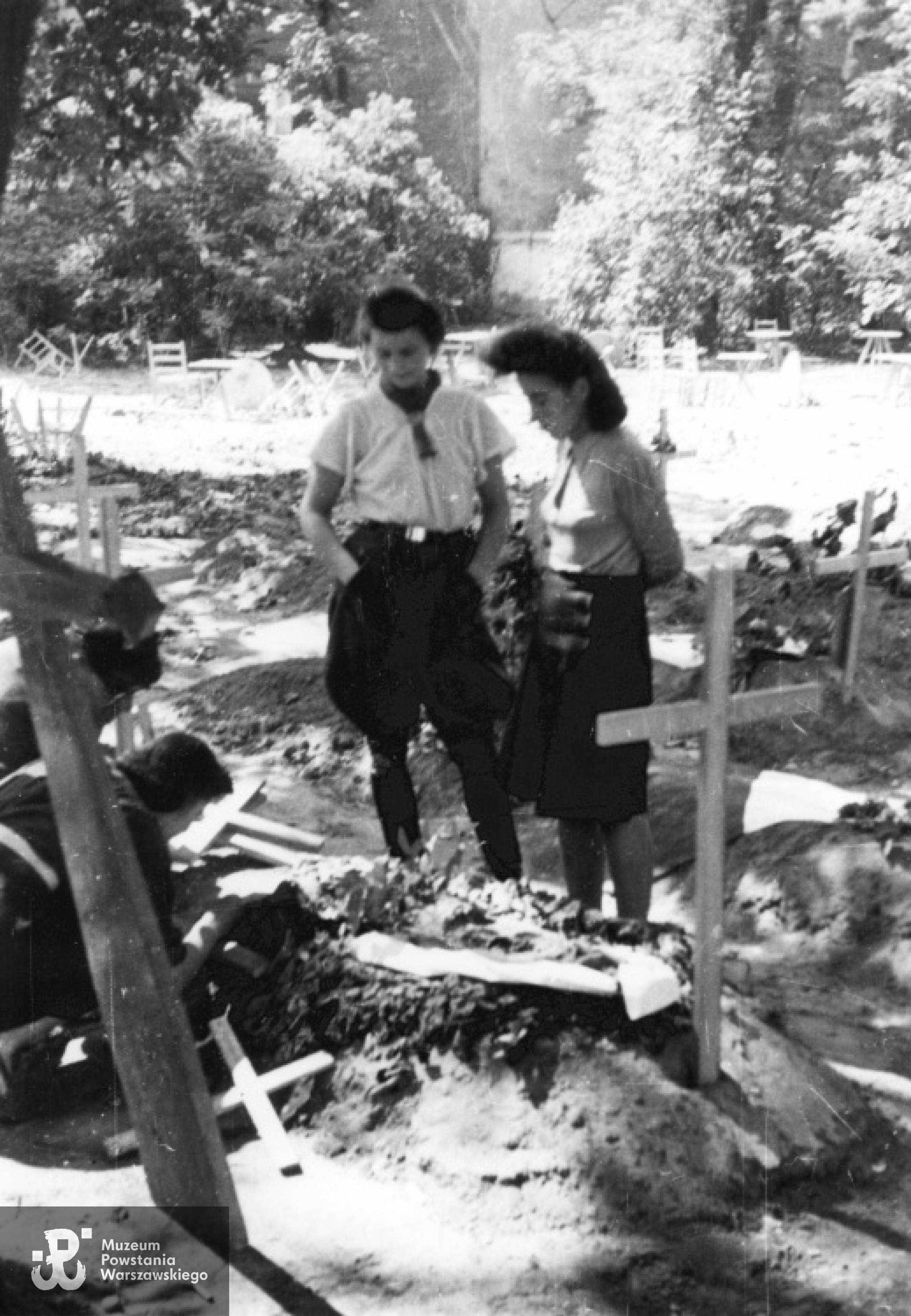 Fotografia z Powstania Warszawskiego autorstwa Eugeniusza Lokajskiego. Łączniczki plutonu 1147 "Rygla" na cmentarzu przy ulicy Mazowieckiej; od lewej: pochylona przy grobie Jadwiga Maydel "Jadwiga", Irena Kummant "Janina", (NN). Ze zbiorów Fototeki MPW, sygn. MPW-IN/260, prawa autorskie MPW.