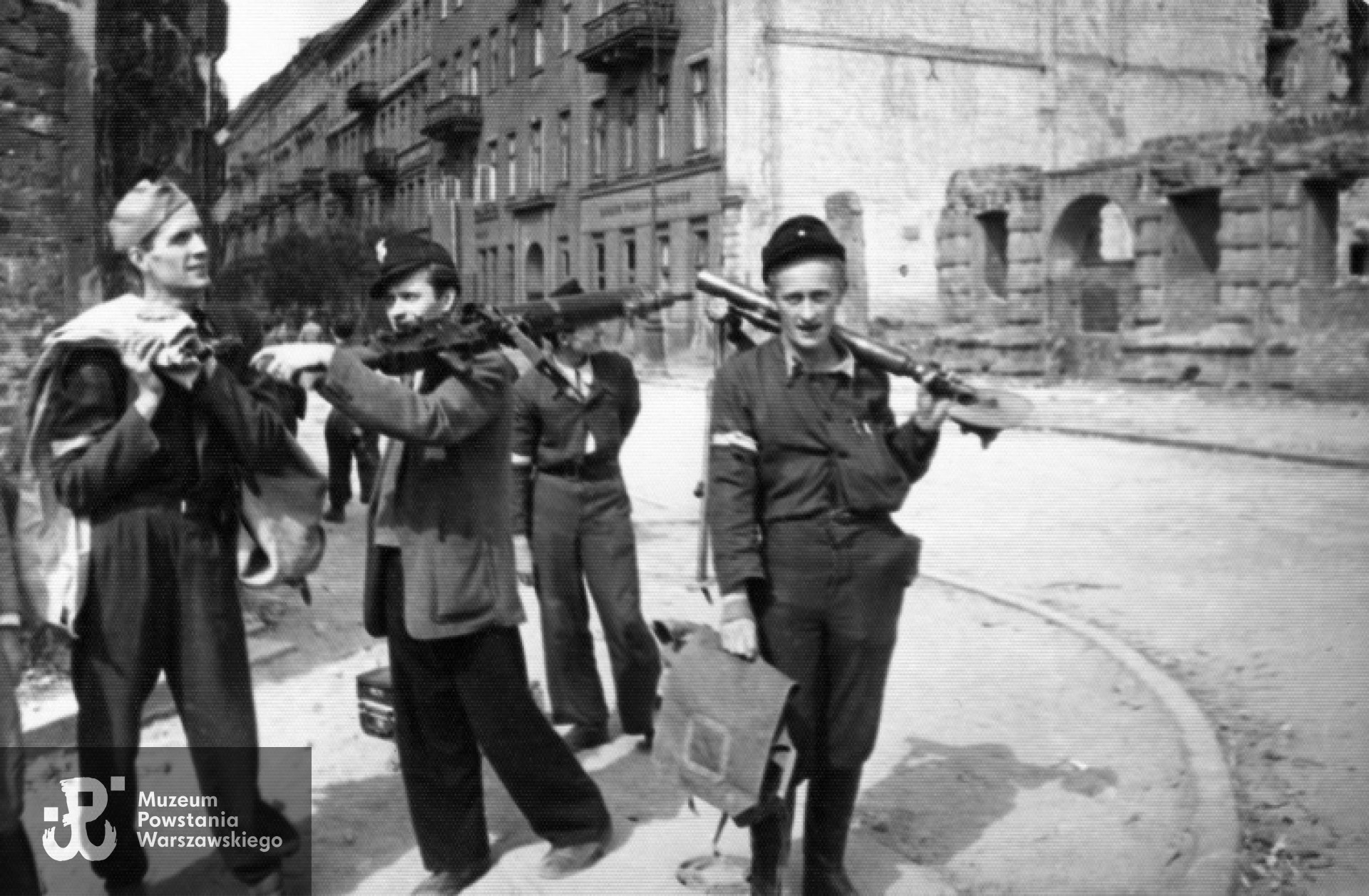 Zdobywcy Komendy Policji obładowani bronią na ul. Czackiego przy Sienkiewicza. Od prawej: rozebrana w czasie okupacji zniszczona kamienica Czackiego nr 2 (róg Świętokrzyska), dalej budynki nr: 4, 6, 8 i 10. Po prawej stoi strz. "Zimny" - Ryszard Lipski z trójnogiem do MG-08/15 Maxim.