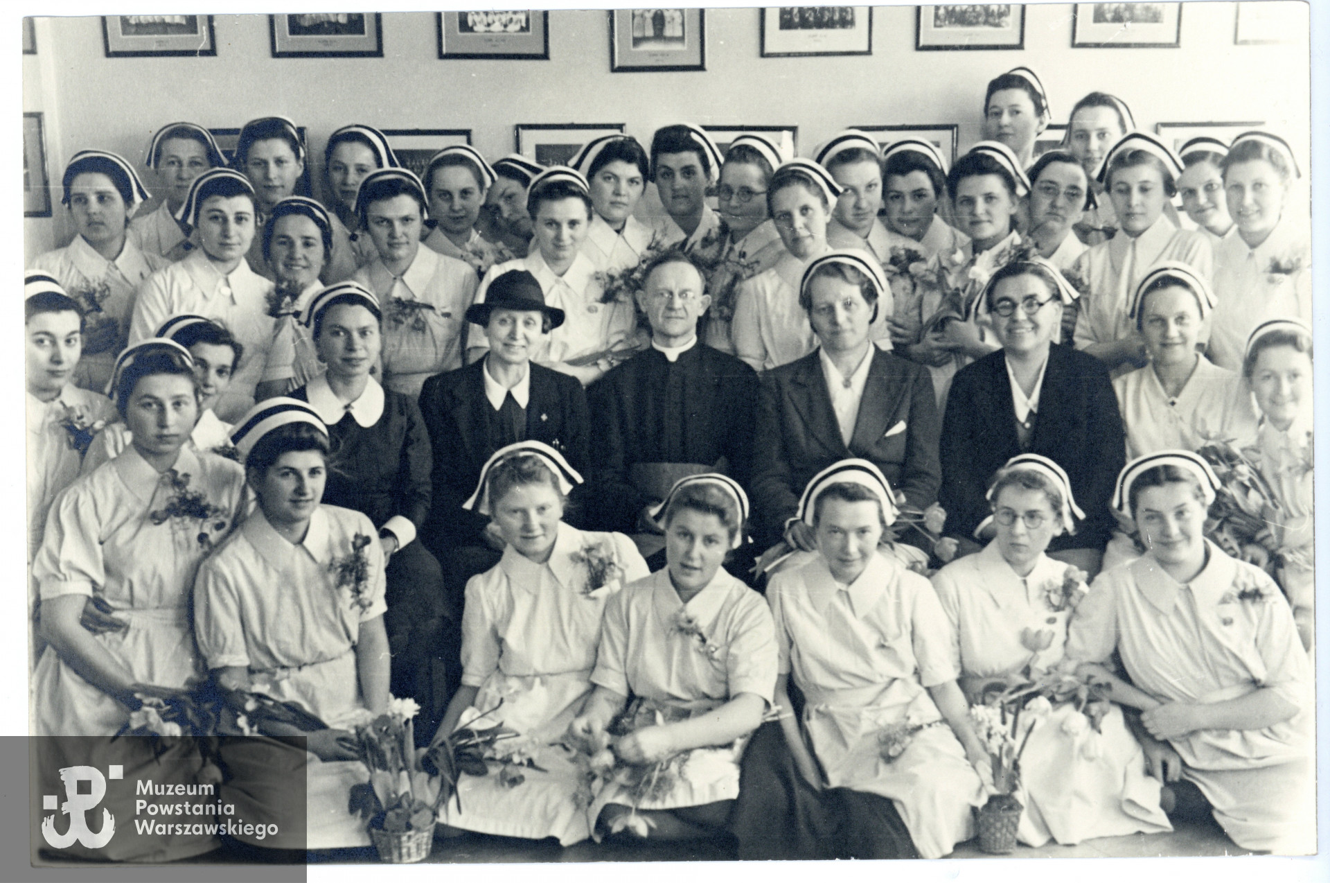 Fotografia przedstawiająca absolwentki Warszawskiej Szkoły Pielęgniarstwa, 1943 rok. W środku grono pedagogiczne, od lewej Małgorzata Żmudzka (1883-1944), przedwojenna instruktorka Warszawskiej Szkoły Pielęgniarstwa, do 1943 roku dyrektorka Szkoły Pielęgniarstwa PCK w Warszawie  na ul. Smolnej 6 (połączonej przez Niemców w 1943 z WSP na Koszykowej 78), ks. płk Tadeusz Julian Jachimowski, naczelny kapelan ZWZ-AK oraz  kapelan Warszawskiej Szkoły Pielęgniarstwa przy ul. Koszykowej 78, obok niego Jadwiga Romanowska, dyrektorka szkoły, następnie wicedyrektorka Wanda Lankajtes. Wśród absolwentek stojących za pedagogami, na lewo od Małgorzaty Żmudzkiej widoczna jest piel. Feliksa Tomaszewska ps. Elipsa. Fot. ze zbiorów rodzinnych, udostępnili p. Jacek Krzysztof Moskwa i p. Stanisława Piotr Moskwa, synowie piel. Feliksy Tomaszewskiej. Skany wykonano w Muzeum Powstania Warszawskiego, 03/2024
