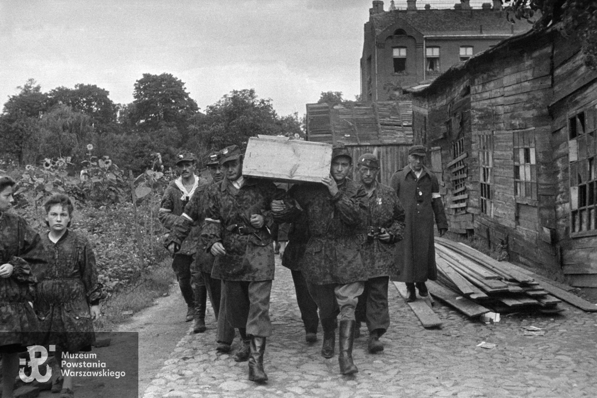  Fotografia z Powstania Warszawskiego. Wola. Żołnierze kompanii "Zemsta" batalionu "Pięść" niosący trumnę poległego ppor. Tadeusza Towarnickiego "Naprawa". Trumnę niosą ppor. Kazimierz Barszczewicz "Henryk" i ppor. NN "Ryś". Z lewej widoczny ppor. Jan Kowalczyk "Kieł", z lornetką po prawej Eugeniusz Kuźwa "Hipek" dowódca 169 plutonu batalionu "Kiliński". W tle po prawej widoczny budynek Zakładu dla umysłowo niedorozwiniętych na posesji Karolkowa 77. Autorem zdjęcia jest Stefan Bałuk. Fot. ze zbiorów Muzeum Powstania Warszawskiego, sygn.   MPW-IS/149. Ujęcie tożsame: MPW-IH/3444.