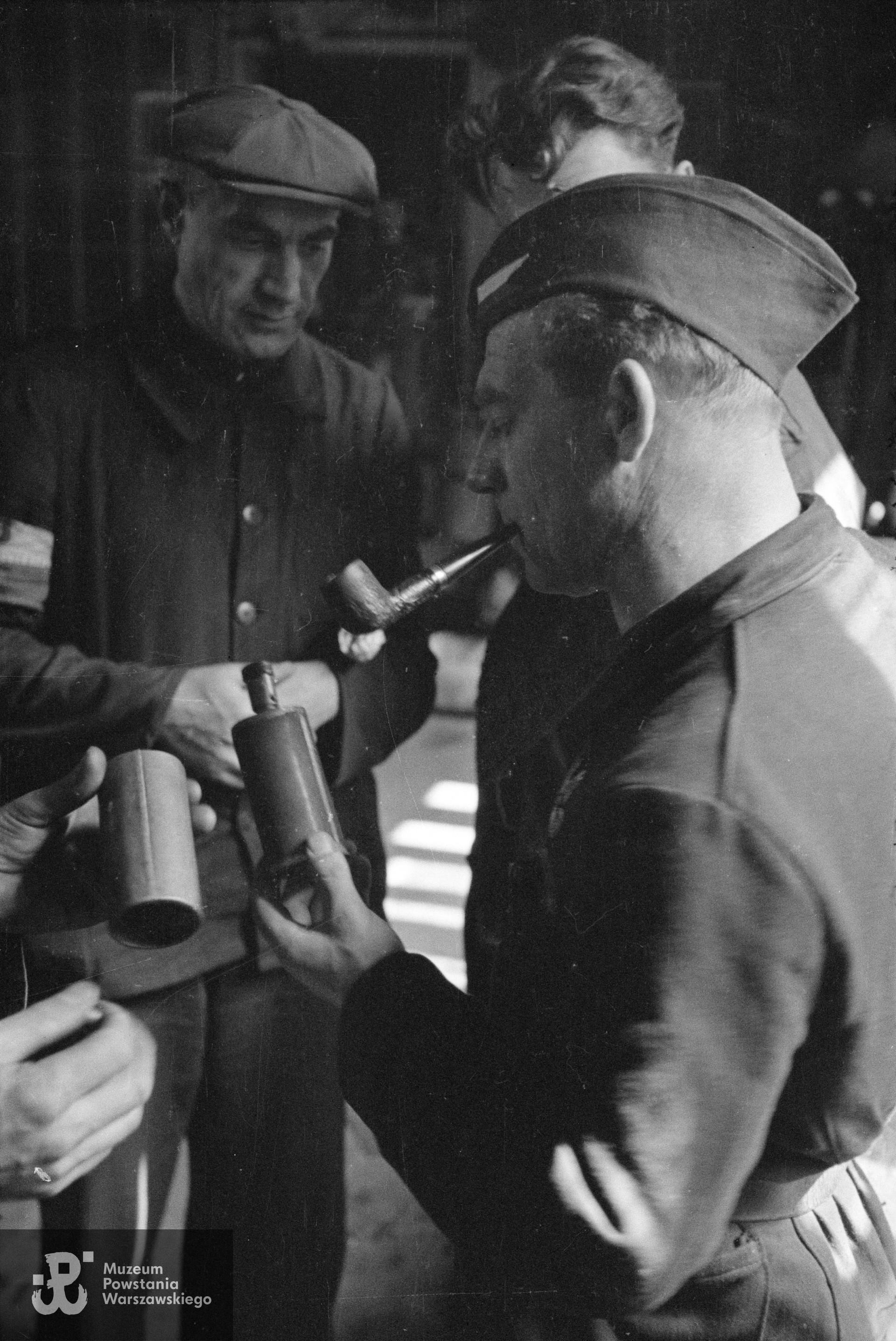 Fotografia z Powstania Warszawskiego. Powiśle. Jedno z pomieszczeń dawnej fabryki Gerlacha przy ul. Tamka 38/40. Por. Stefan Berent "Steb" (na pierwszym planie z fajką) oglądający "sidolówkę" - granat zaczepny powstańczej produkcji, którego obudowę stanowiły blaszane pojemniki od płynu do czyszczenia metali kolorowych o handlowej nazwie Sidol. Na drugim planie po lewej st. wachm. Roch Kafarowski "Ksawery".   Fot. ze zbiorów Muzeum Powstania Warszawskiego, sygn. MPW-IN/692, autorem zdjęcia jest Eugeniusz Lokajski "Brok". 