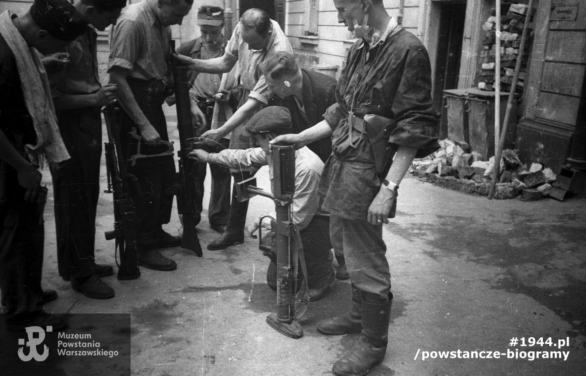 Fotografia z Powstania Warszawskiego. Śródmieście Północne. Podwórze kamienicy przy ul. Świętokrzyskiej 20. Plut. pchor. Adam Podkóliński "Michał", "Olszewski" (trzeci od lewej) prezentuje grupie powstańców z kompanii "Janusza" z batalionu "Harnaś" niemiecki lekki karabin maszynowy (leMG 08/15) zdobyty po ataku na Komendę Policji. Powstaniec na pierwszym planie po prawej trzyma brytyjski granatnik przeciwpancerny PIAT. Fot. ze zbiorów Muzeum Powstania Warszawskiego, sygn. MPW-IN/10203. Autorem zdjęcia jest Joachim Joachimczyk "Joachim"
