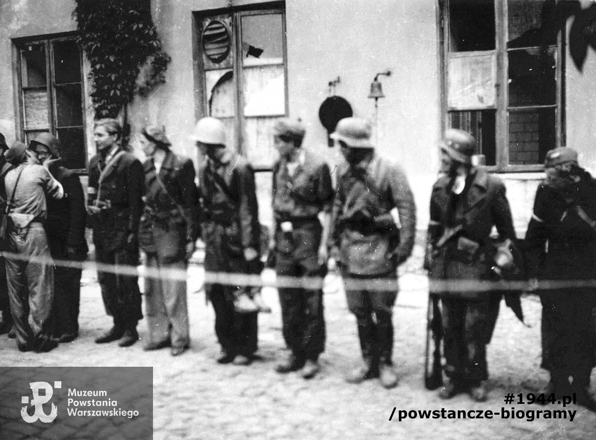 Fotografia z Powstania Warszawskiego. Zbiórka powstańców z plutonu "Rygla" zgrupowania "Bartkiewicz" po zakończonej akcji w podwórzu zdobytej Komendy Policji. Po lewej st. strz. Zbigniewa Kurbiela "Krakusa" całuje mjr Bernard Romanowski "Wola". Reszta żołnierzy w szeregu, od lewej: pierwszy strz. Wiesław Nałęcz-Mroczkowski "Wiesiek", drugi Józef Korczyński "Józef", trzeci kpr. pchor. Kazimierz Jesionowski "Jesion", IV, V i VI żołnierz nierozpoznani, siódmy od lewej stoi  strz. Mieczysław Śliwiński "Mietek". Fot. ze zbiorów Muzeum Powstania Warszawskiego, sygn. MPW-IP/3829
