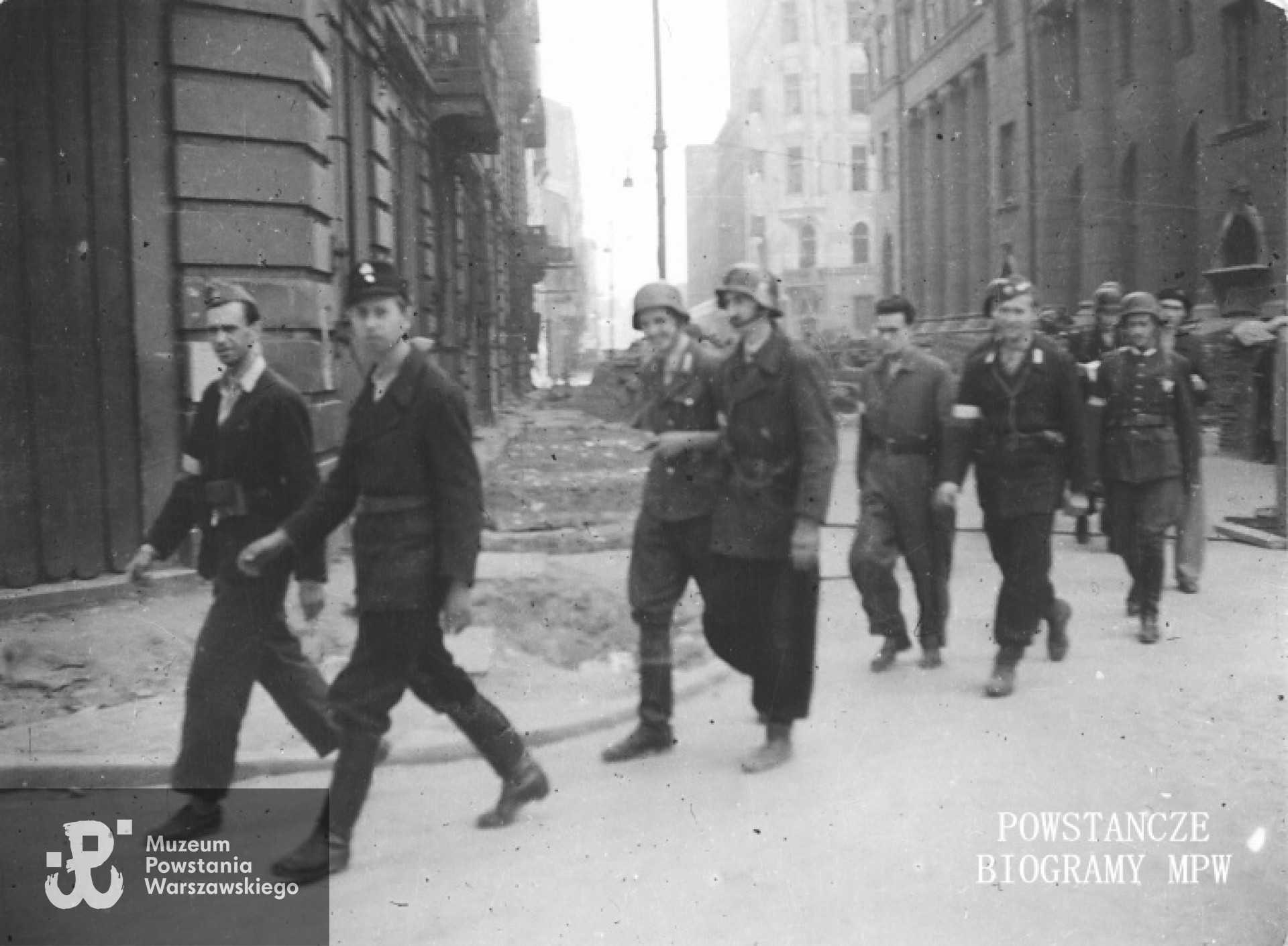 Fotografia z Powstania Warszawskiego. Idący dwójkami powstańcy z  plutonu "Rygla" zgrupowania "Bartkiewicz", skręcający z ul. Świętokrzyskiej w Szkolną, ujęcie w kierunku wsch. Idą od lewej: kpr. z cenz. Jerzym Jenike "Zwierz", kpr. pchor. Janusz Sawicki "Zabawa", kpr. pchor. Andrzej Gliński "Leszczyc", strz. Władysław Mydell "Waldemar" st. strz. Zbigniew Kurbiel "Krakus", kpr. pchor. Andrzej Strawiński "Chimera". Na wprost wylot Świętokrzyskiej. Po lewej na pierwszym planie kamienica Świętokrzyska 36/38/40 róg Jasna 11 róg Szkolna 2. Po prawej gmach PKO Świętokrzyska 31/33 róg Jasna 9, dalej widoczny narożnik kamienicy Świętokrzyska 27 róg Jasna 16. Autor zdjęcia Irena Kummant-Skotnicka "Janina". MPW - sygn. MPW-IP/3845