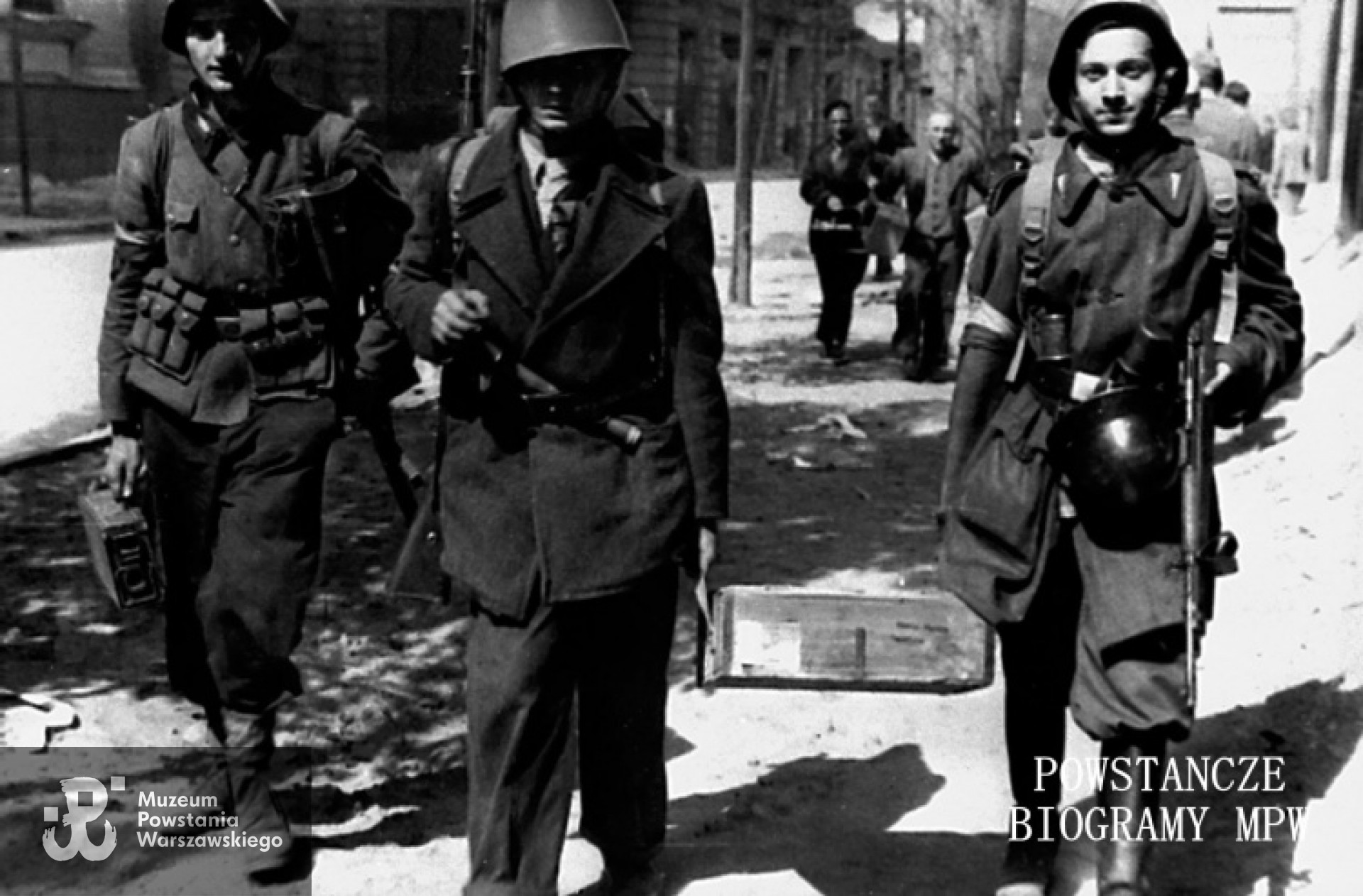 Fotografia z Powstania Warszawskiego. Śródmieście Północne. Powstańcy niosący skrzynki z amunicją na ulicy Jasnej nieopodal skrzyżowania ze Świętokrzyską. Ujęcie w kierunku północnym. W tle po lewej widoczny fragment gmachu PKO - Jasna 9 róg Świętokrzyska 31/33. Pierwszy po prawej stronie idzie kpr. Zbigniew Olszewski "Sęp" z kompanii "Koszta". Zdjęcie wykonane po zdobyciu broni i amunicji w Komendzie Policji przy Krakowskim Przedmieściu 1. Fot. ze zbiorów Muzeum Powstania Warszawskiego, sygn. MPW-IH/4324