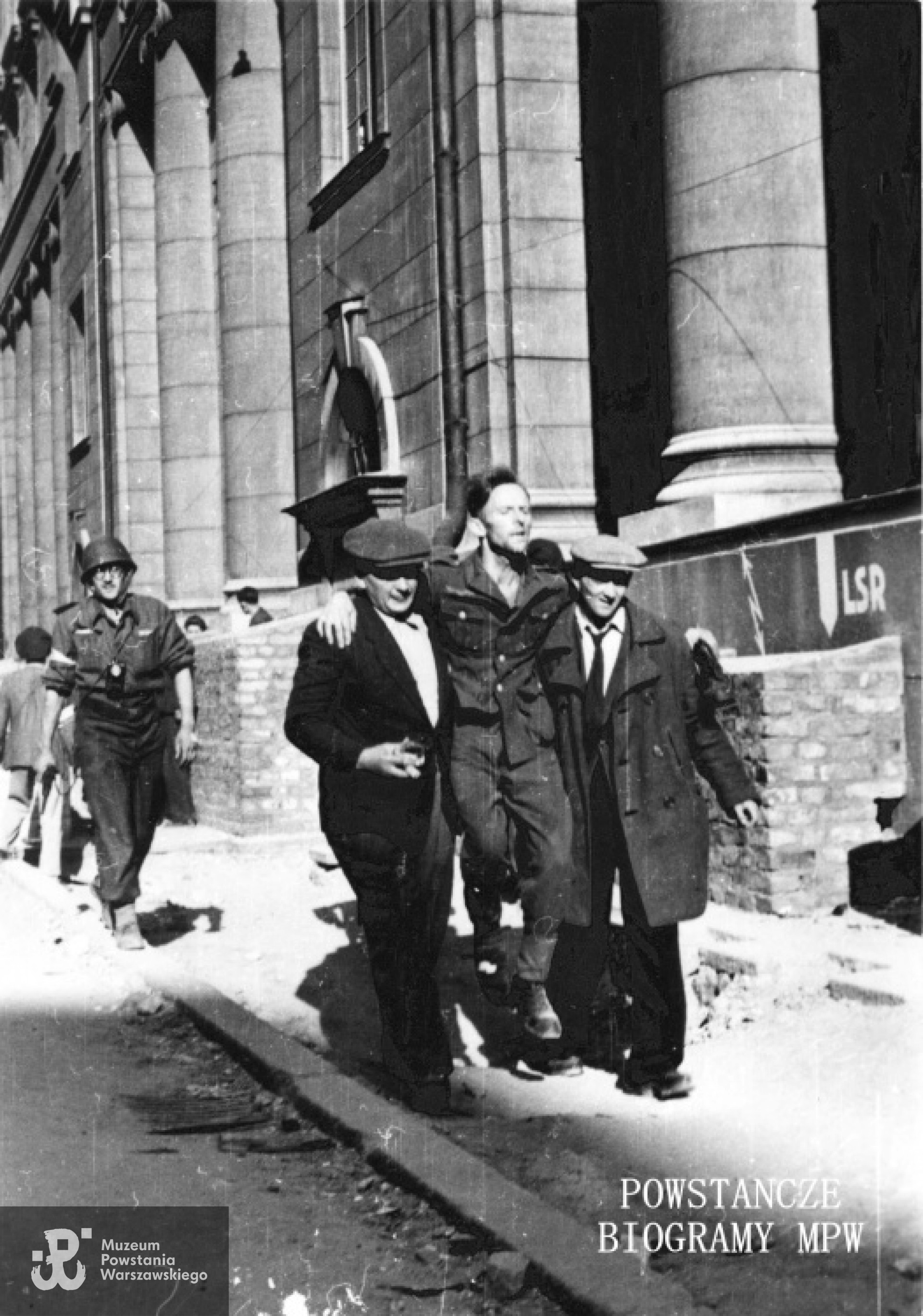 Fotografia z Powstania Warszawskiego. Śródmieście Północne, ul. Świętokrzyska. Ranny żołnierz niemiecki przenoszony przez dwóch cywili, pod eskortą uzbrojonego powstańca, ze zbombardowanego obozu jenieckiego w pobliżu hotelu "Victoria" do gmachu PKO. Z tyłu najprawdopodobniej  Witold Stanisławski "Rola" (w hełmie, z latarką wykorzystywaną przy przejściu kanałami na szyi.) Zdjęcie autorstwa Eugeniusza Lokajskiego, ze zbiorów Fototeki MPW, sygn. <a href=" http://www.1944.pl/galerie/fototeka/foto/242/?szukaj=lokajski"> MPW-IN/116</a>
