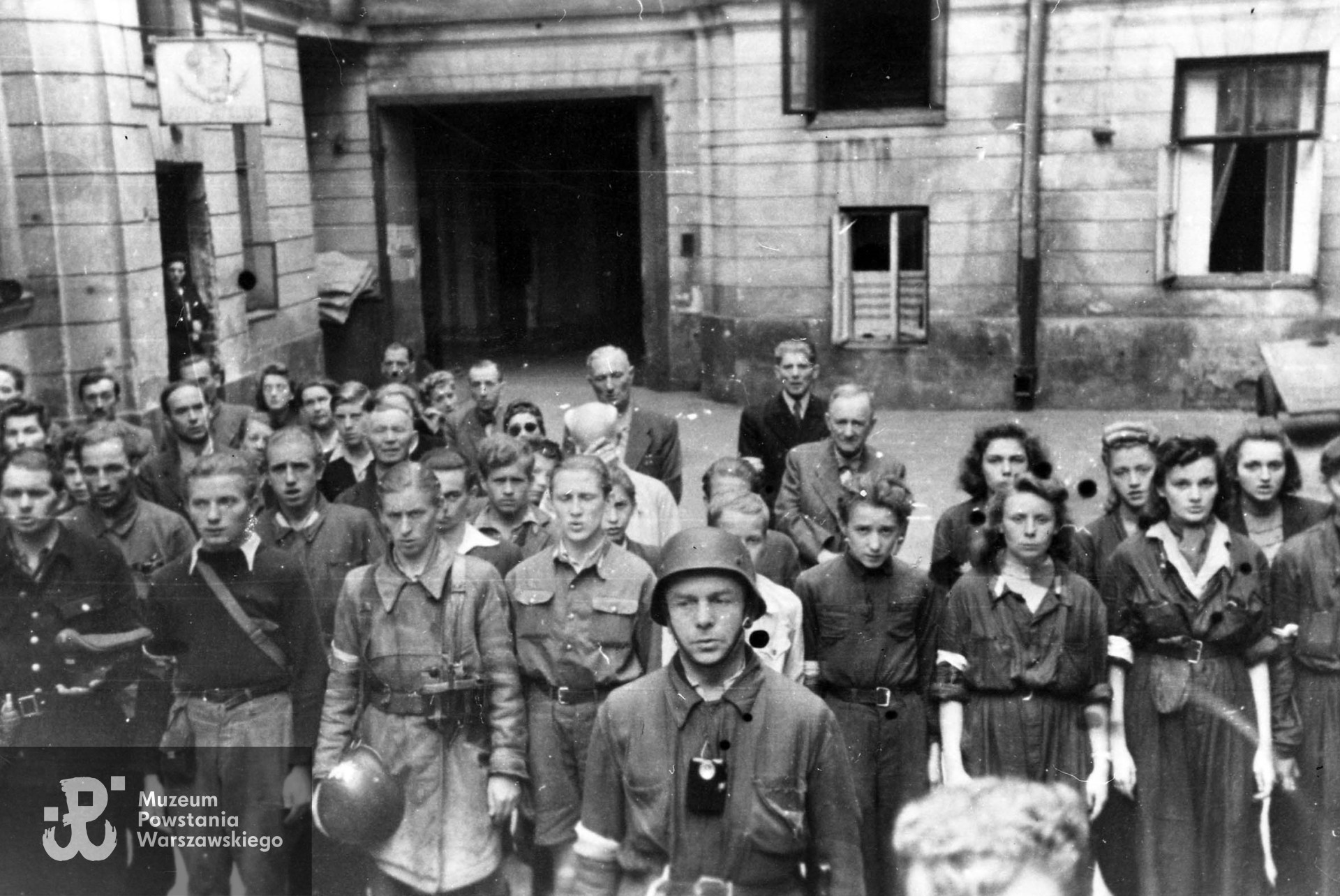 Fotografia z Powstania Warszawskiego. Śródmieście Południowe, rejon pl. Trzech Krzyży. Grupa powstańców z 1. plutonu "Kłosa" 3. kompanii "Redy" batalionu "Miłosz" podczas mszy św. na podwórzu kamienicy. Autorem zdjęcia jest Wojciech Marczyński "Bogusław". Fot. ze zbiorów Muzeum Powstania Warszawskiego, sygn. MPW-IH/101