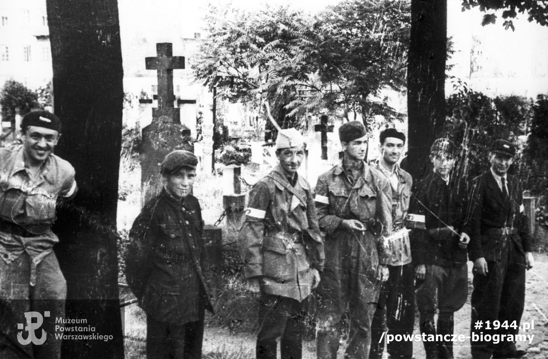 Fotografia z Powstania Warszawskiego. Wola. Godzina "W". Zbiórka powstańców z plutonu "Agaton" Batalionu "Pięść" na terenie cmentarza ewangelicko-augsburskiego. Od lewej: st. strz. Kazimierz Piechotka "Jacek", (NN), kpt. Stanisław Jankowski "Agaton", strz. Piotr Woyzbun "Janusz", strz. Zygmunt Skrobański "Blikle", kpr. pchor. Artur Czarnomski "Kleofas" i kpr. Seweryn Brwiliński "Adam". W tle po prawej widoczne zabudowania przy ul. Mireckiego z narożną kamienicą przy ul. Okopowej 39. Autorem zdjęcia jest Stefan Bałuk "Starba", "Kubuś".  Fot. ze zbiorów Muzeum Powstania Warszawskiego, sygn. MPW-IH/3418