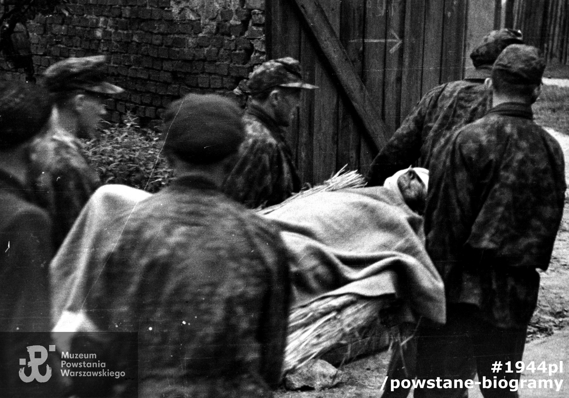 Fotografia z Powstania Warszawskiego. Wola. Ewakuacja rannego kpr. pchor. Adama Grymaszewskiego "Jastrząb" z kompanii "Zemsta" z Batalionu "Pięść" w rejonie cmentarza ewangelicko-augsburskiego. Nad noszami, w środkowej części fotografii, widoczny z profilu kpr. pchor. Stefan Rogowiec "Szaruga", ze zbiorów Muzeum Powstania Warszawskiego MPW-IK/1292