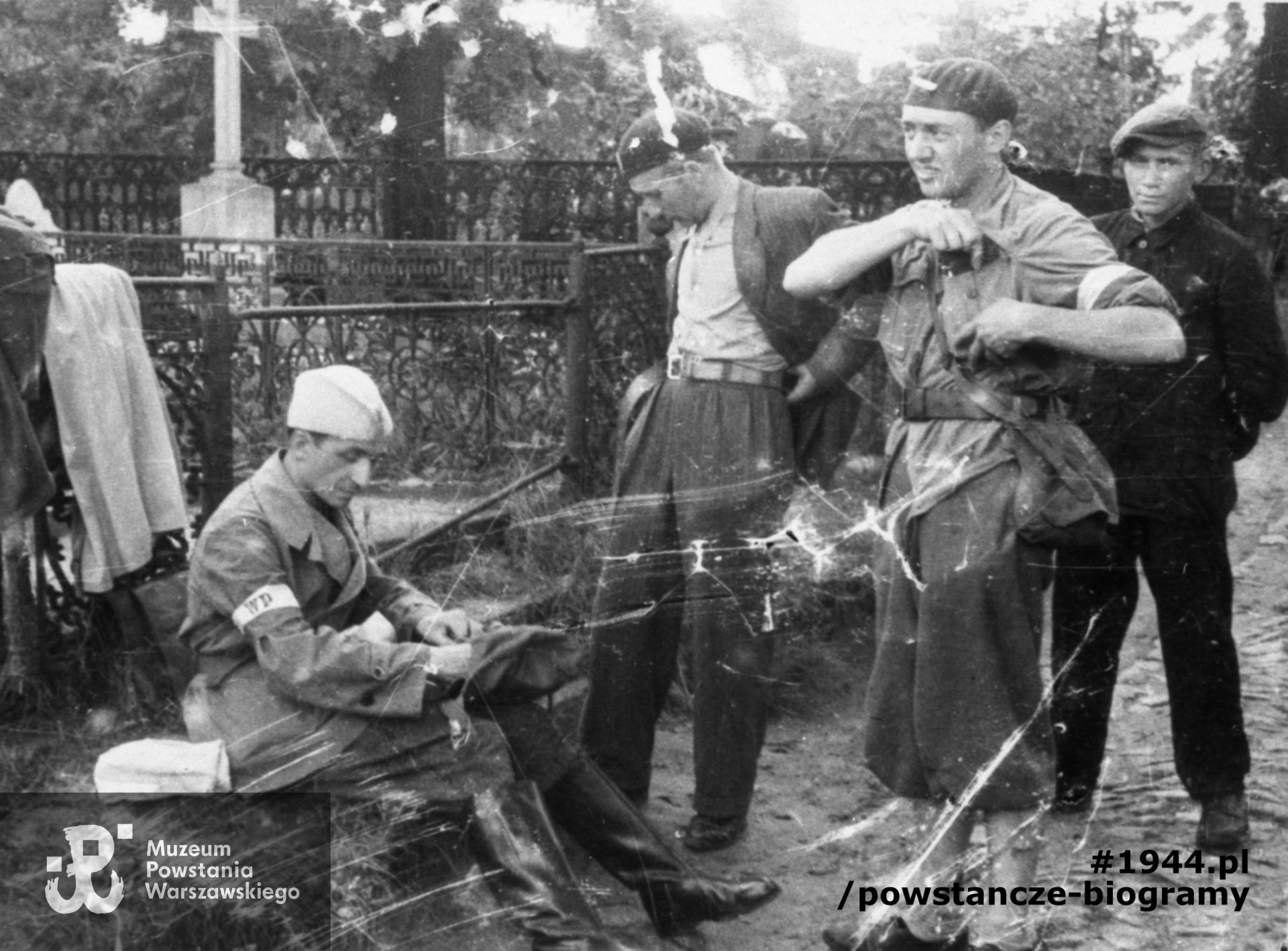 Fotografia z Powstania Warszawskiego. Wola. Godzina "W". Koncentracja powstańców z plutonu "Agaton" Batalionu "Pięść" na terenie cmentarza ewangelicko-augsburskiego, w rejonie bramy od ul. Karolkowej. Od lewej: siedzi kpt. Stanisław Jankowski "Agaton", kpr. Czesław Wegner "Bartek", st. strz. Kazimierz Piechotka "Jacek", (NN). W tle widoczny grobowiec rodziny Baumgart (z białym krzyżem). Autorem zdjęcia jest Stefan  Bałuk. Fot. ze zbiorów  Muzeum Powstania Warszawskiego, sygn. MPW-IH/2164