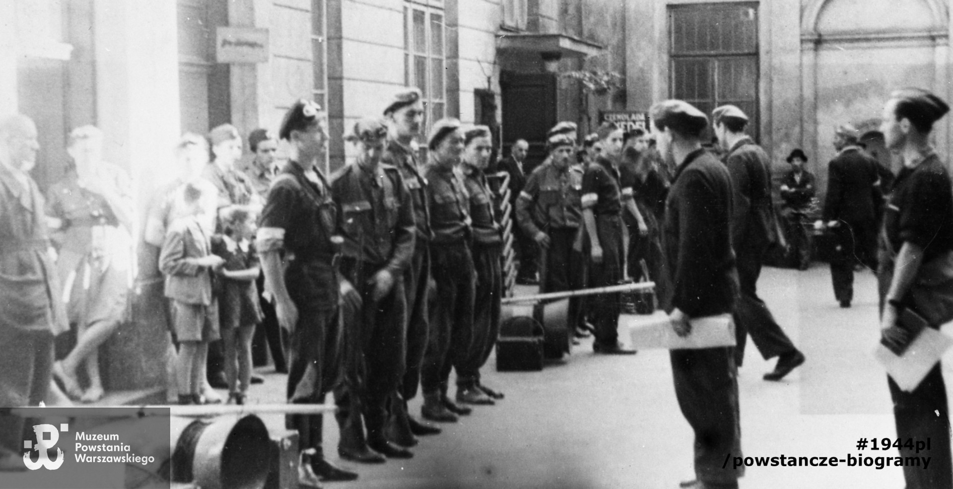 Fotografia z Powstania Warszawskiego. Śródmieście Północne. Odprawa oddziału tzw. "megafoniarzy" na podwórzu domu Wedla przy ul. Szpitalnej 8. Pierwszy od lewej Janusz Kulesza ps. "Super", dalej NN ps. "Wicher", Feliks Lendzion ps "Wołg". Fot. ze zbiorów Muzeum Powstania Warszawskiego, sygn. MPW-IH/4879