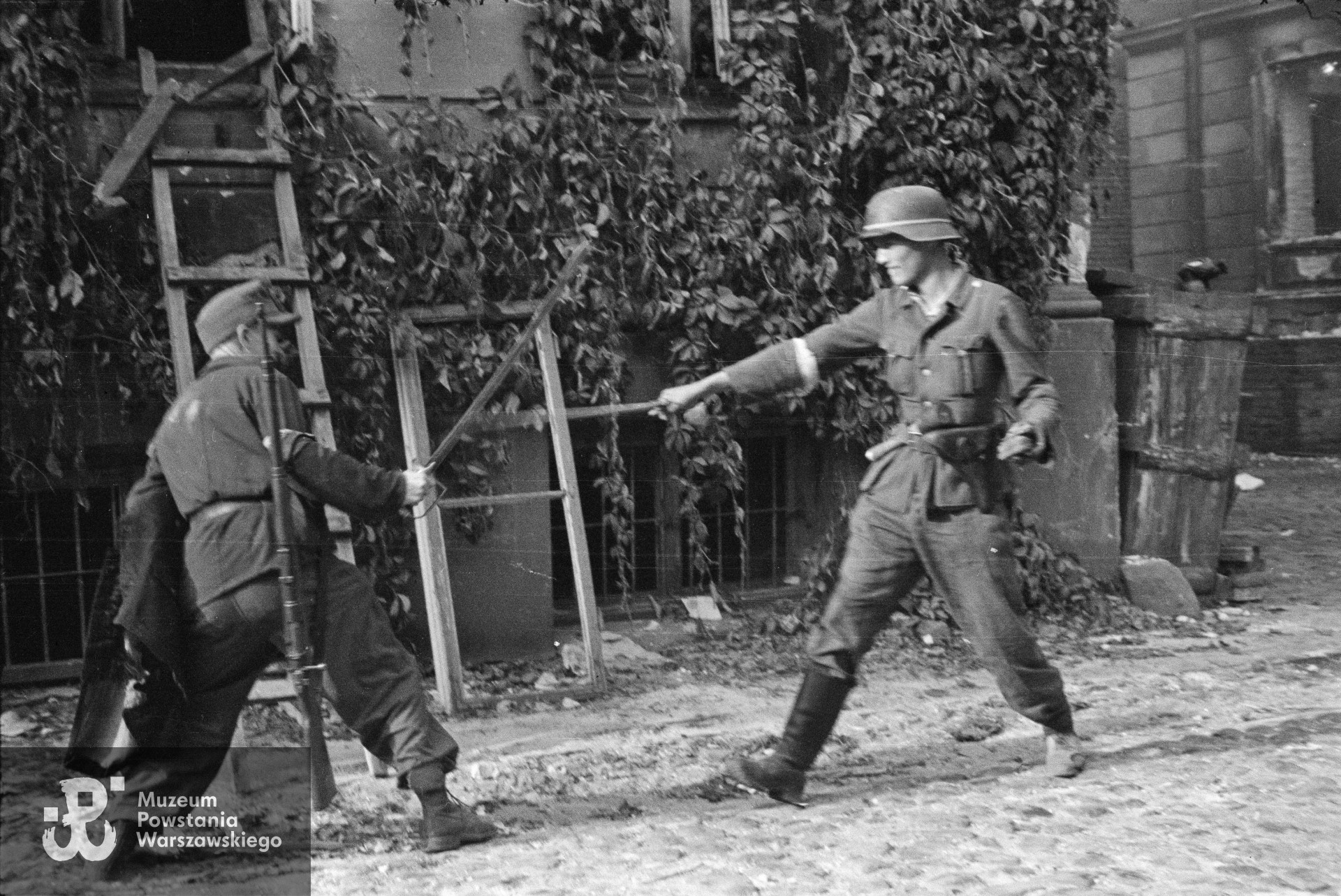 Fotografia z Powstania Warszawskiego: Śródmieście Północne - 23 sierpnia 1944 r.   Inscenizowany pojedynek na szable dwóch powstańców z kompanii "Koszta" na dziedzińcu zdobytego gmachu Komendy Policji przy Krakowskim Przedmieściu 1. Po prawej plut. pchor. Jerzy Tyczyński "Jur". Fot. autorstwa Eugeniusza Lokajskiego ps. "Brok". Ze zbiorów Muzeum Powstania Warszawskiego, sygn. MPW-IN/754.