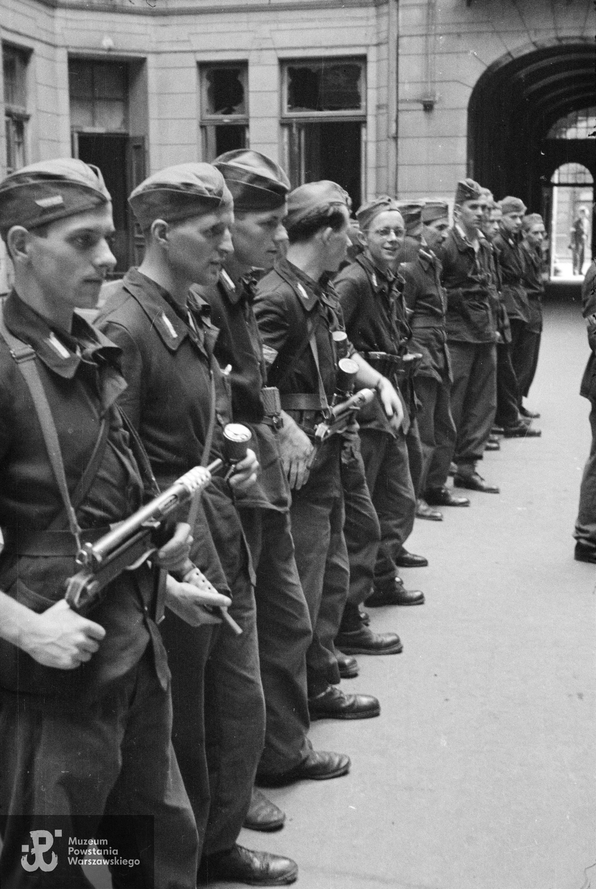 Fotografia z Powstania Warszawskiego. Śródmieście Północne. Apel III plutonu Oddziału Osłonowego Wojskowych Zakładów Wydawniczych (WZW) przy Oddziale VI Sztabu Komendy Głównej AK na podwórzu kamienicy Lewenfisza przy pl. Napoleona 1, róg ul. Boduena 2, róg ul. Sienkiewicza 1. Ujęcie w kierunku południowym. W tle widoczna brama wychodząca na ulicę Boduena. Stoją od lewej: ppor. Józef Kulesza "Mały Dżo", ppor. Władysław Wojtkiewicz "Ohm", st. strz. Jerzy Słubicki "Jarema", kpr. pchor. Tadeusz Bruszewski "Sztubak", kpr. Roman Cichalewski "Romek”, „Okularnik”, (NN), (NN), (NN), (NN), strz. Włodzimierz Leo "Bawół" i kpr. pchor. Ryszard Wrzosek "Dick". Autorem zdjęcia jest Eugeniusz Lokajski "Brok". Fot. ze zbiorów Muzeum Powstania Warszawskiego, sygn. MPW-IN/715