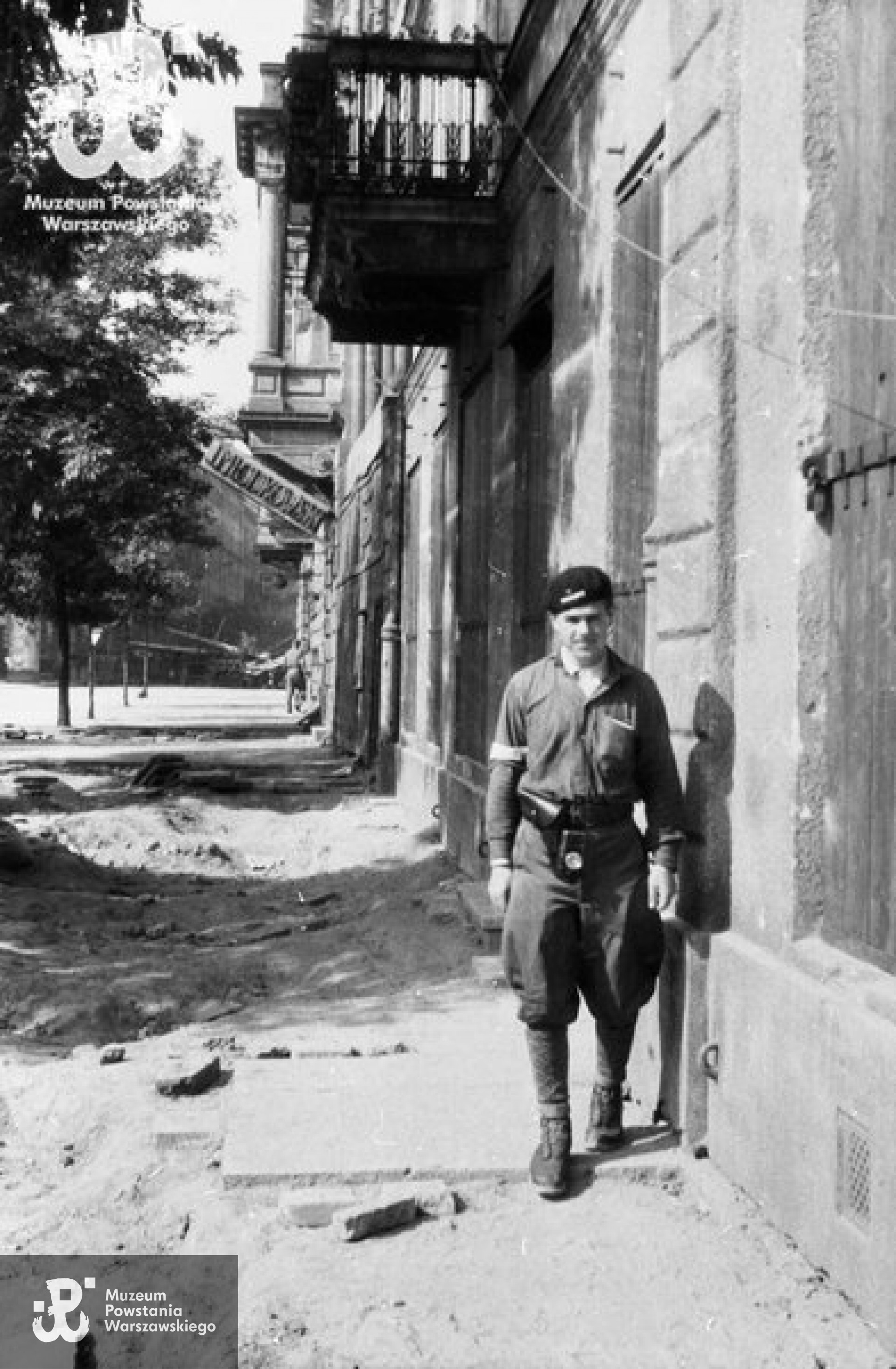 Fotografia z Powstania Warszawskiego. Ppor. Stanisław Sasinowski "Żbik", dowódca 3 plutonu II kompanii szturmowej batalionu OW - KB "Sokół" przy kamienicy pl. Trzech Krzyży 16. Widoczny neon Magazynu Kapeluszy Męskich J. Młodkowski w kamienicy pl. Trzech Krzyży 18. W głębi widoczna barykada przy wylocie ul. Żurawiej. Fot. autorstwa Mariana Grabskiego ps. „Wyrwa“, ze zbiorów Fototeki MPW, sygn. <a href=" http://www.1944.pl/galerie/fototeka/foto/3454/?szukaj=sasinowski"> MPW-IN/1407 </a>