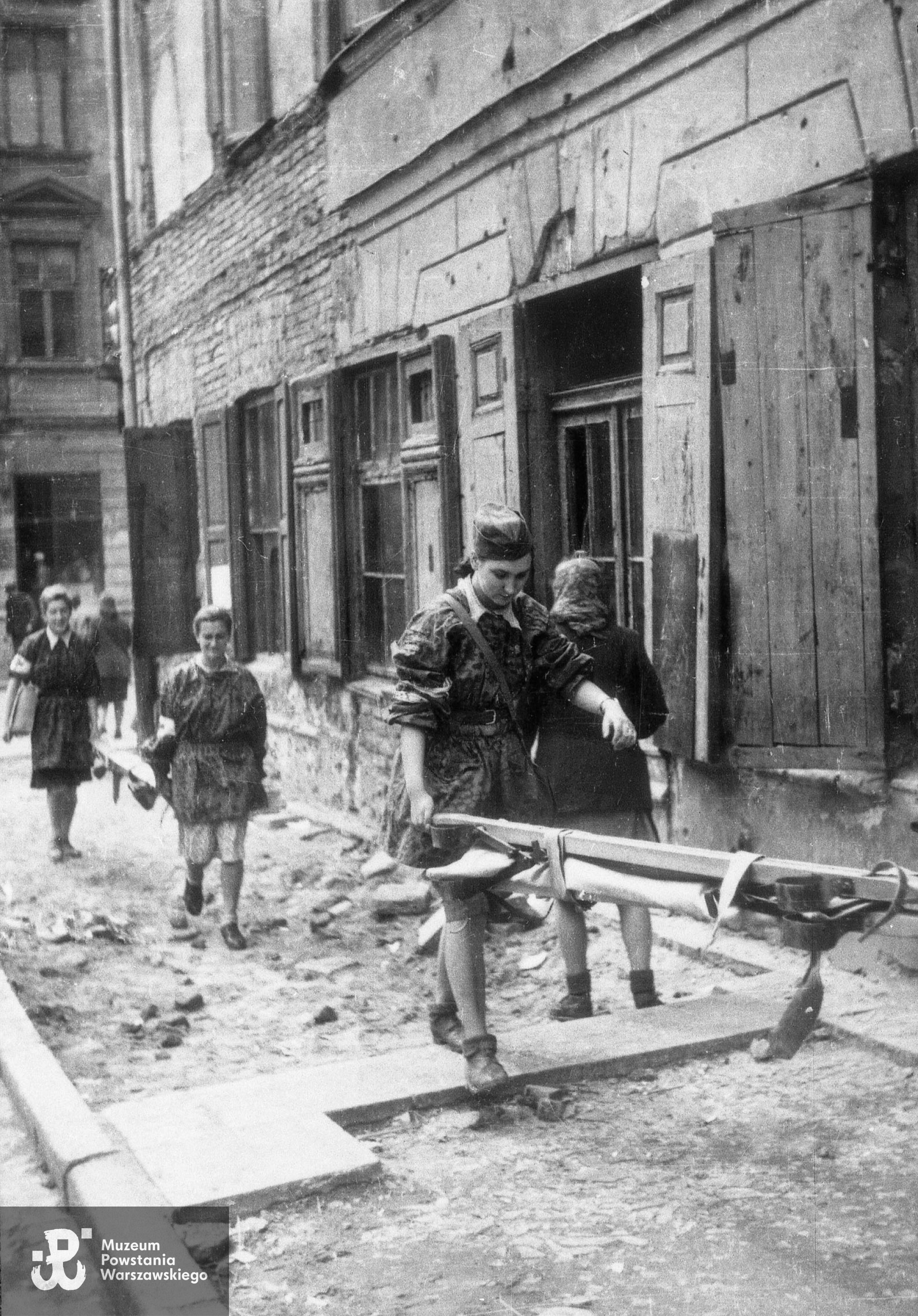 Fotografia z Powstania Warszawskiego. Stare Miasto. Patrol sanitarny przy ul. Piekarskiej, ujęcie w kierunku Podwala. W tle po prawej kamienica Piekarska 22, w głębi po lewej Podwale 11 róg Kapitulnej 1. Autorem zdjęcia jest Jerzy Chojnacki "Chojna". Zdjęcie ze zbiorów Muzeum Powstania Warszawskiego, sygn. MPW-IH/2234. Według informacji przekazanych przez wnuka Wiesławy Kamper-Gliszczyńskiej przypuszczalnie pierwszą z widocznych na zdjęciu dziewcząt  jest "Sławka".