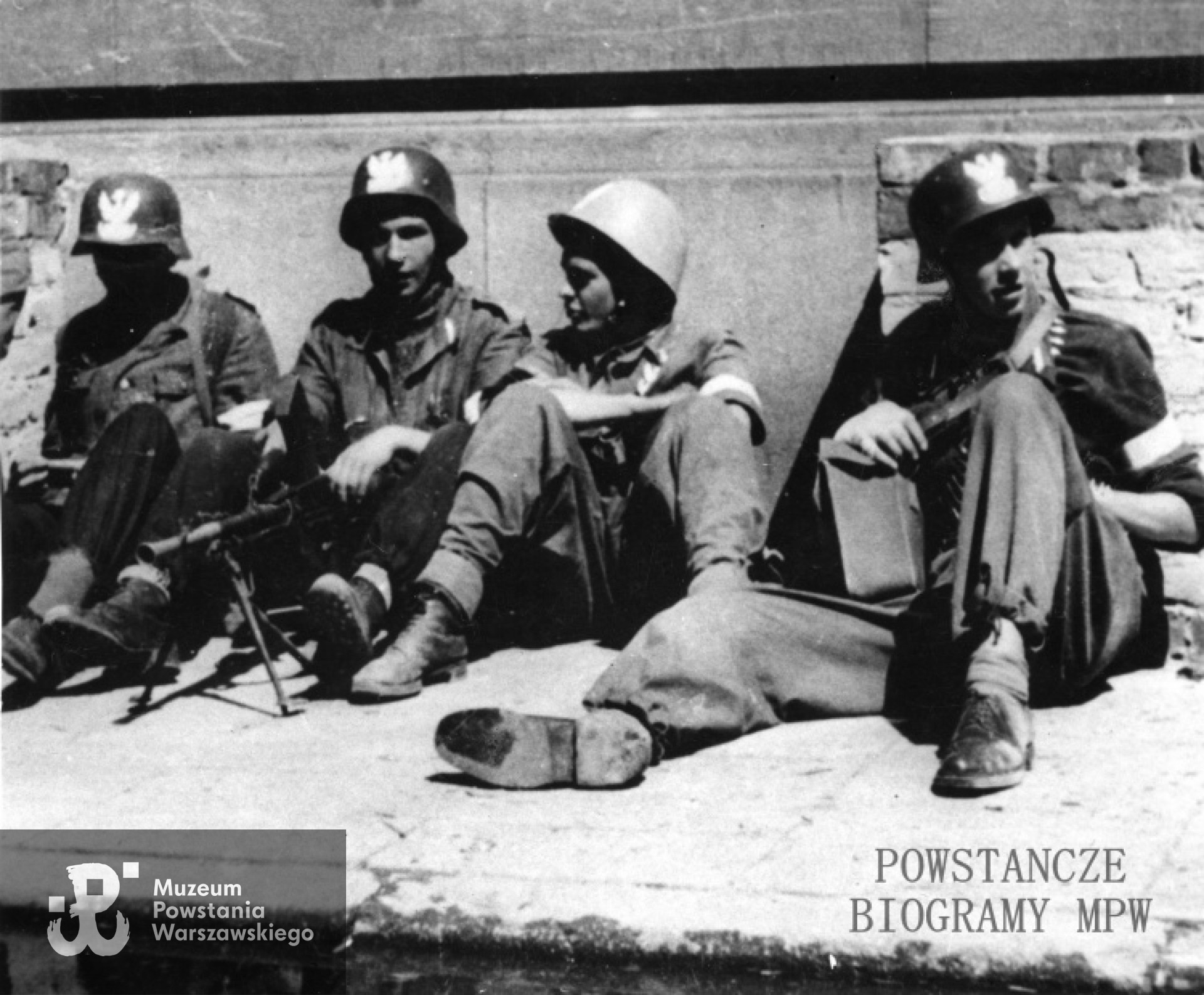 Fotografia z Powstania Warszawskiego. Powiśle. Powstańcy z plutonu "Rafałków" Zgrupowania "Konrad" odpoczywający na chodniku przy wjeździe na dziedziniec gmachu ZNP przy ul. Smulikowskiego 6/8. Kadr większego ujęcia. Jerzy Lech Morawski "Maks" siedzi drugi od lewej. Fot. ze zbiorów prywatnych.