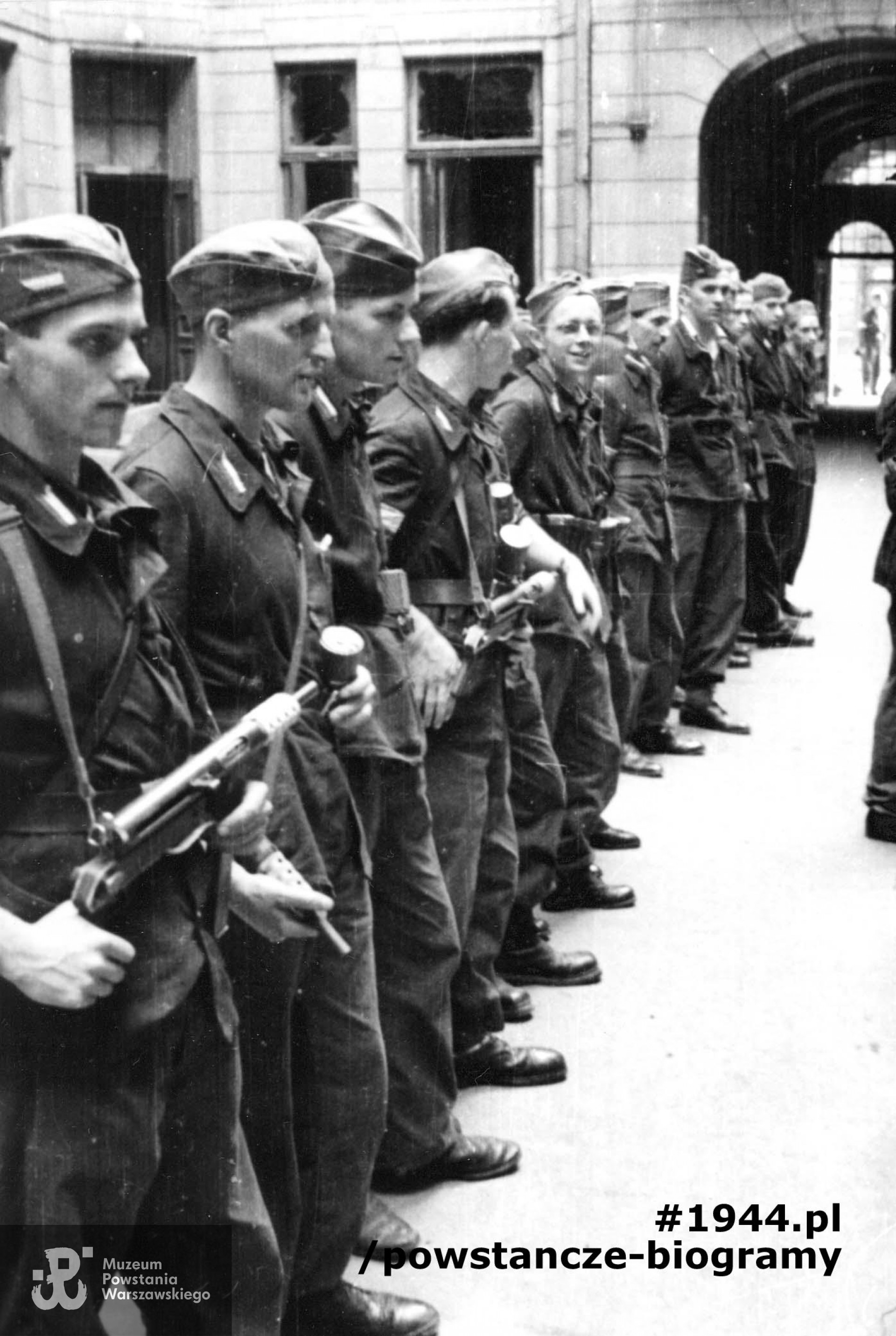 Fotografia z Powstania Warszawskiego. Śródmieście Północne. Apel III plutonu Oddziału Osłonowego Wojskowych Zakładów Wydawniczych (WZW) przy Oddziale VI Sztabu Komendy Głównej AK na podwórzu kamienicy Lewenfisza przy pl. Napoleona 1, róg ul. Boduena 2, róg ul. Sienkiewicza 1. Ujęcie w kierunku południowym. W tle widoczna brama wychodząca na ulicę Boduena. Stoją od lewej: ppor. Józef Kulesza "Mały Dżo", ppor. Władysław Wojtkiewicz "Ohm", st. strz. Jerzy Słubicki "Jarema", kpr. pchor. Tadeusz Bruszewski "Sztubak", kpr. Roman Cichalewski "Romek”, „Okularnik”, (NN), (NN), (NN), (NN), strz. Włodzimierz Leo "Bawół" i kpr. pchor. Ryszard Wrzosek "Dick". Autorem zdjęcia jest Eugeniusz Lokajski "Brok". Fot. ze zbiorów Muzeum Powstania Warszawskiego, sygn. MPW-IN/715