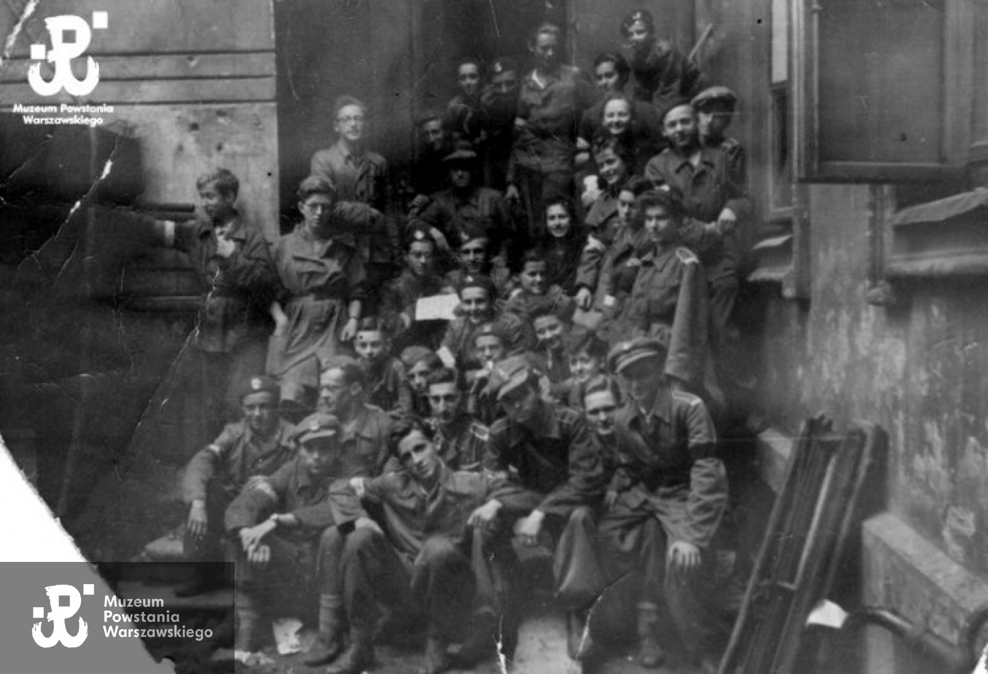 Fotografia z Powstania Warszawskiego. Śródmieście Północne, ulica Chmielna. Zdjęcie grupowe 2 Harcerskiej Baterii Artylerii Przeciwlotniczej „Żbik” Zgrupowanie „Gurt” przed wejściem do Zakładów Graficznych B. Wierzbicki i Spółka w podwórzu budynku przy ulicy Chmielnej 61.Osoby widoczne na zdjęciu: kan. Zbigniew Martyński „Gruda”, kpr. z cenz. Marcin Czerwiński „Smugoń”, kan. Robert Skupieński „Kretowicz”, kpr. z cenz. Jerzy Bieńkowski „Rudzki”, bomb. Zygmunt Rakowiecki „Rola”, kan. Marcin Lewandowicz „Mosiądz”, plut. z cenz. Stefan Bogdanowicz „Malicki”, z-ca d-cy plut. I, plut. z cenz. Tadeusz Błażyński „Ładosz”, „Świda”, kan. łączn. Wacław Makarczyk „Sokół”, kan. łączn. Andrzej Dąbrowski „Tygrys”, kpr. pchor. Piort Osiński „Kaczanowski”, d-ca plut. II, łączn. NN "Orska", strz. sanit. Krystyna Kamać „Szafrańska”, kan. Andrzej Czałbowski „Kruk”, z-ca d-cy SOA, strz. łączn. Maria Sturgólewska „Poręba”, kan. łączn. Tomasz Wołowski „Sum”, st. strz. łączn. Irena Glass „Kim”, kan. Aleksander Lubarski „Dryświaty - Modrzewski”, kan. Kazimierz Rakowski „Świteź" a>, sanit. Stanisława Gawlik „Stenia”, strz. łączn. Barbara Danuta Reinhard „Tancred”, kpr. Elżbieta Tatarkiewicz „Żabska”, st. strz. Janusz Eysymontt „Szczerbina”, NN „Roman” z ŻOB, poczet dowódcy, strz. Aleksander Bober „Alosza”, „Babinicz", Gruzin, były kapitan Armii Czerwonej, k-tka łączn. Halina Loga „Średzka”, kpr. sanit. Maria Jezierska „Bogdańska” , k-tka sanitariuszek, kan. Adam Bromke „Karski”, kan. anusz Preyss „Kowalski”, kan. łączn. Ryszard Boreński „Ryś”, bomb. Wojciech Psarski „Nałęcz”, kpr. z cenz. Marian Łączyński „Orlik” , z-ca d-cy plut. II, st. strz. łączn. Lucyna Piotrowska „Boj”, kan. Witold Janczewski „Sowa”. Fot. Joachim Joachimczyk. Ze zbiorów Fototeki MPW, sygn. <a href=" http://www.1944.pl/galerie/fototeka/foto/7397/autor/63/pageNumber/63/"> MPW-IP/2110</a>