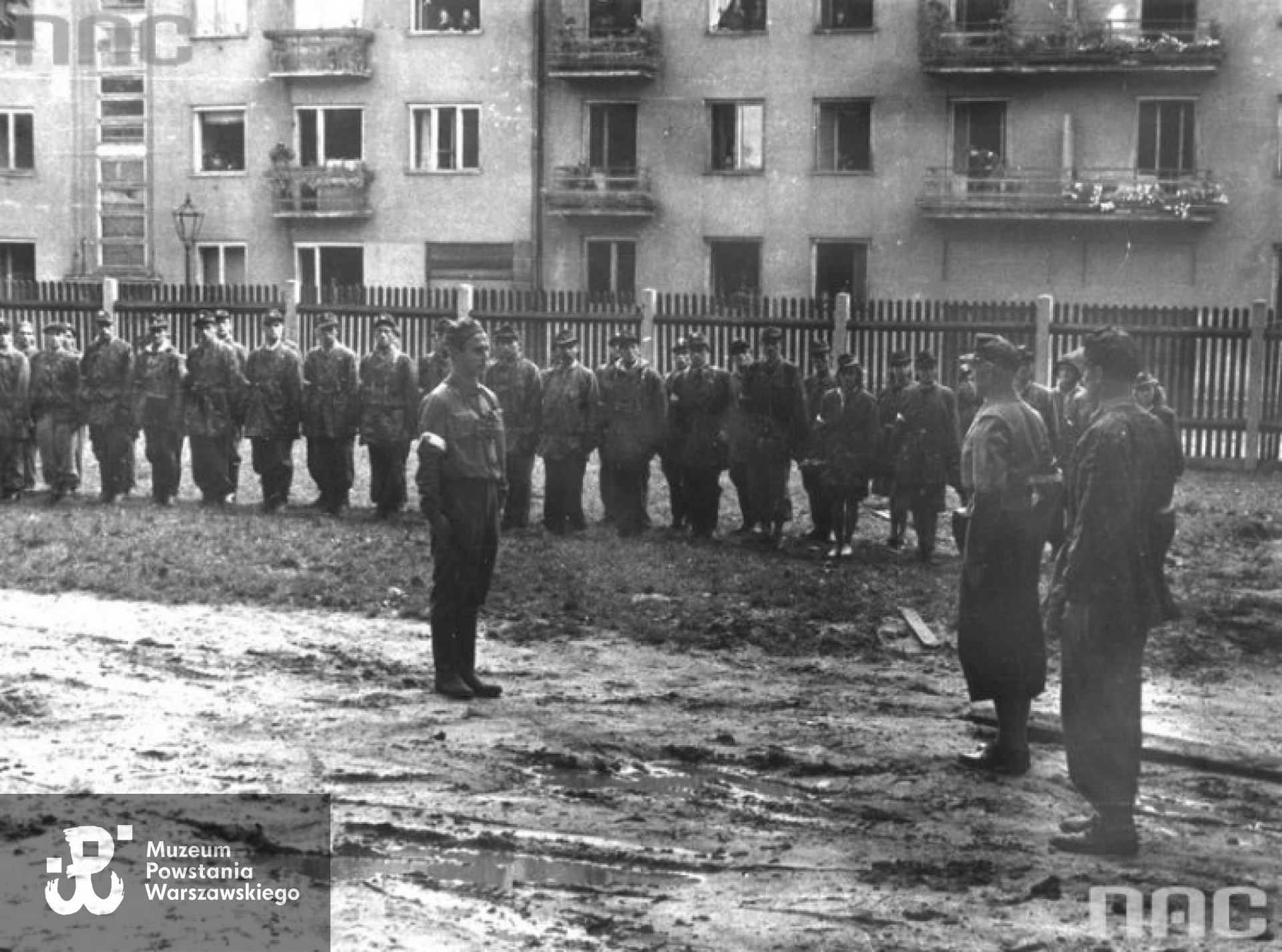 Powstanie Warszawskie: por. Stefan Matuszczyk ps. "Porawa" składa meldunek dowódcy batalionu mjr. Alfonsowi Kotowskiemu ps. "Okoń". W tle widoczni żołnierze III plutonu i kobiecy patrol sanitarny kompanii, ul. Karolkowa (za ogrodzeniem) i kamienica Karolkowa 80. <i> [www.audiovis.nac.gov.pl] </i>