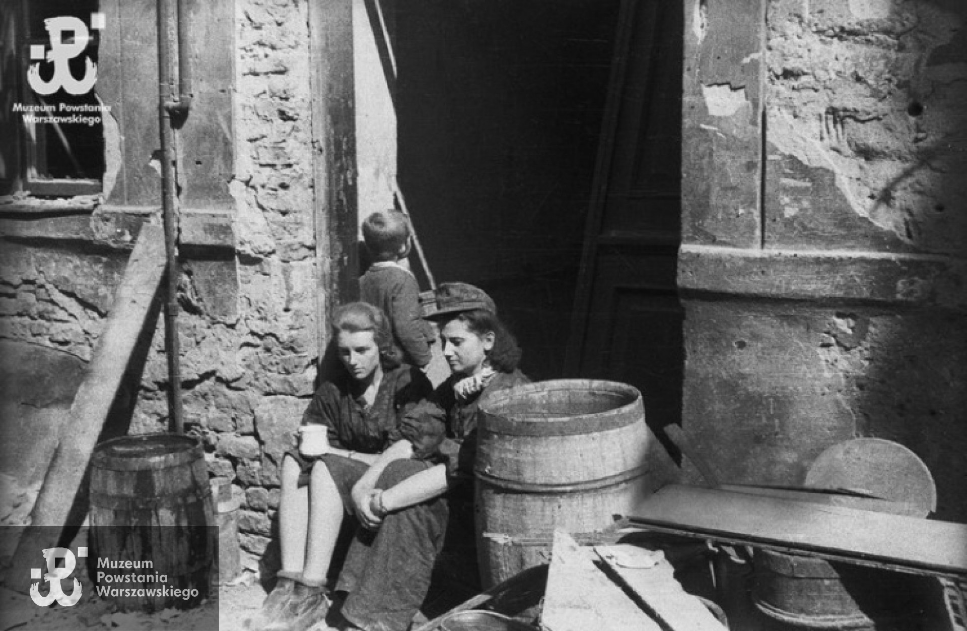 Powstanie Warszawskie, sierpień 1944 - Stare Miasto. Sanitariuszki z kompanii "Anna” batalionu "Gustaw”: Danuta Magreczyńska "Danka” (po lewej) i Wanda Bagniewska "Wanda” na progu domu przy ulicy Ślepej. Za nimi chłopiec, jeden z mieszkańców kamienicy ukrywających się w piwnicach. Fot. Wiesław Chrzanowski "Wiesław", zbiory Fototeki MPW, sygn. MPW-IN/3245