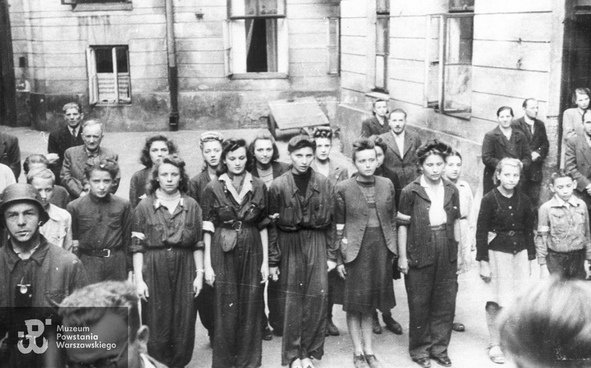 Fotografia z Powstania Warszawskiego. Śródmieście Południowe. Sanitariuszki i łączniczki z 1. plutonu „Kłosa” 3. kompanii „Redy” batalionu „Miłosz” podczas mszy św. odprawianej na podwórzu kamienicy przy ul. Mokotowskiej.  W pierwszym rzędzie stoją od lewej: w hełmie Ryszard Lewandowski „Oporek”, za nim nieco zasłonięty łącznik Ziuk (w jasnej kurtce), łącznik NN, obok niego Wacława Sutowska ps. Aćka,   Hanna Baranowska „Lalka”, Krystyna Kleszczewska „Krysia”, patrolowa „Teresa” (prawdopodobnie  Zofia Tomasik), Janina Kleszczewska „Kopernik„, NN, NN. W drugim rzędzie stoją: za „Aćką” - „Dzidka” (prawdopodobnie Helena Schabińska), obok niej Daniela Białowąs „Niutka” (za H. Baranowską „ Lalką"), następnie "Jadwiga", NN i "Alusia" (prawdopodobnie Alicja  Gilewska). Pozostałe osoby nierozpoznane. Autorem zdjęcia jest Wojciech Marczyński ”Bogusław„. Fot. ze zbiorów Muzeum Powstania Warszawskiego, sygn. MPW-IH/5916 