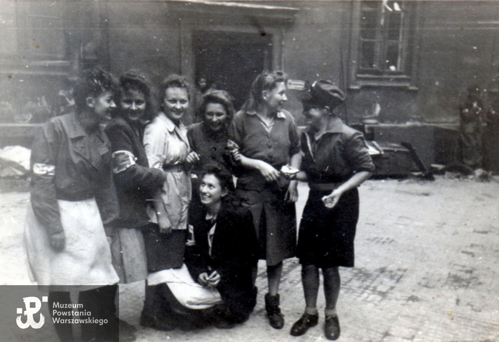 Fotografia z Powstania Warszawskiego autorstwa Joachima Joachimczyka ps. "Joachim".  Śródmieście Północne. Grupa sanitariuszek i łączniczek z plutonu 569 -  2. Harcerskiej Baterii Artylerii Przeciwlotniczej "Żbik" na podwórzu kamienicy Chmielna 57. Na zdjęciu od lewej: NN, sanit. Krystyna Kamać "Szafrańska", sanit. Jadwiga Mundkowska "Dina Pękałówna",  łączn. Irena Loga "Modrzewska", łączn. Halina Loga "Średzka", łączn. Maria Sturgólewska "Poręba",sanit. Stanisława Gawlik "Stenia". Ze zbiorów Muzeum Powstania Warszawskiego, sygn. MPW-IP/2109