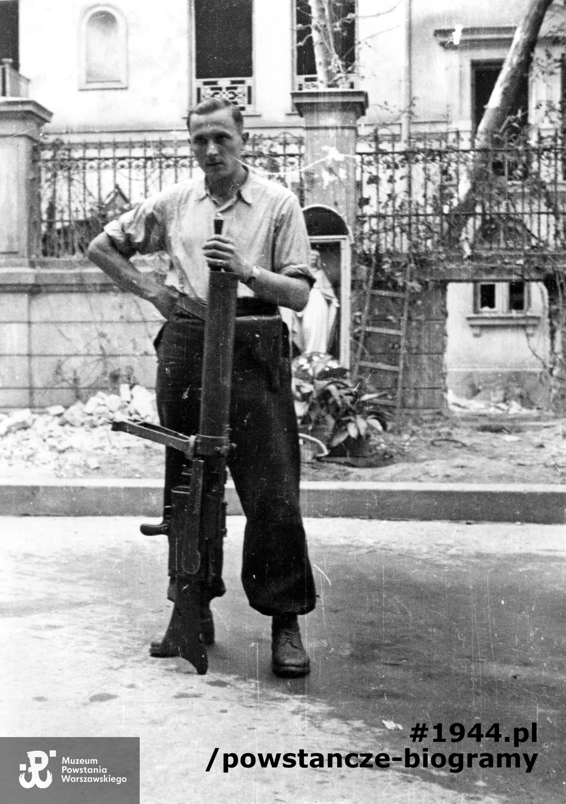 Fotografia z Powstania Warszawskiego. Plut. pchor. Adam Podkóliński "Michał" z kompanii "Janusz", batalionu "Harnaś" pozuje ze zdobytym po ataku na Komendę Policji niemieckim karabinem maszynowym na podwórzy kamienicy przy ul. Świętokrzyskiej 20. W tle widoczna kapliczka, a za ogrodzeniem fragment elewacji bocznej (południowej) oficyny domu na posesji przy ul. Mazowieckiej 4. Autorem zdjęcia jest Eugeniusz Lokajski "Brok". Fot. ze zbiorów Muzeum Powstania Warszawskiego, sygn. MPW-IN/731