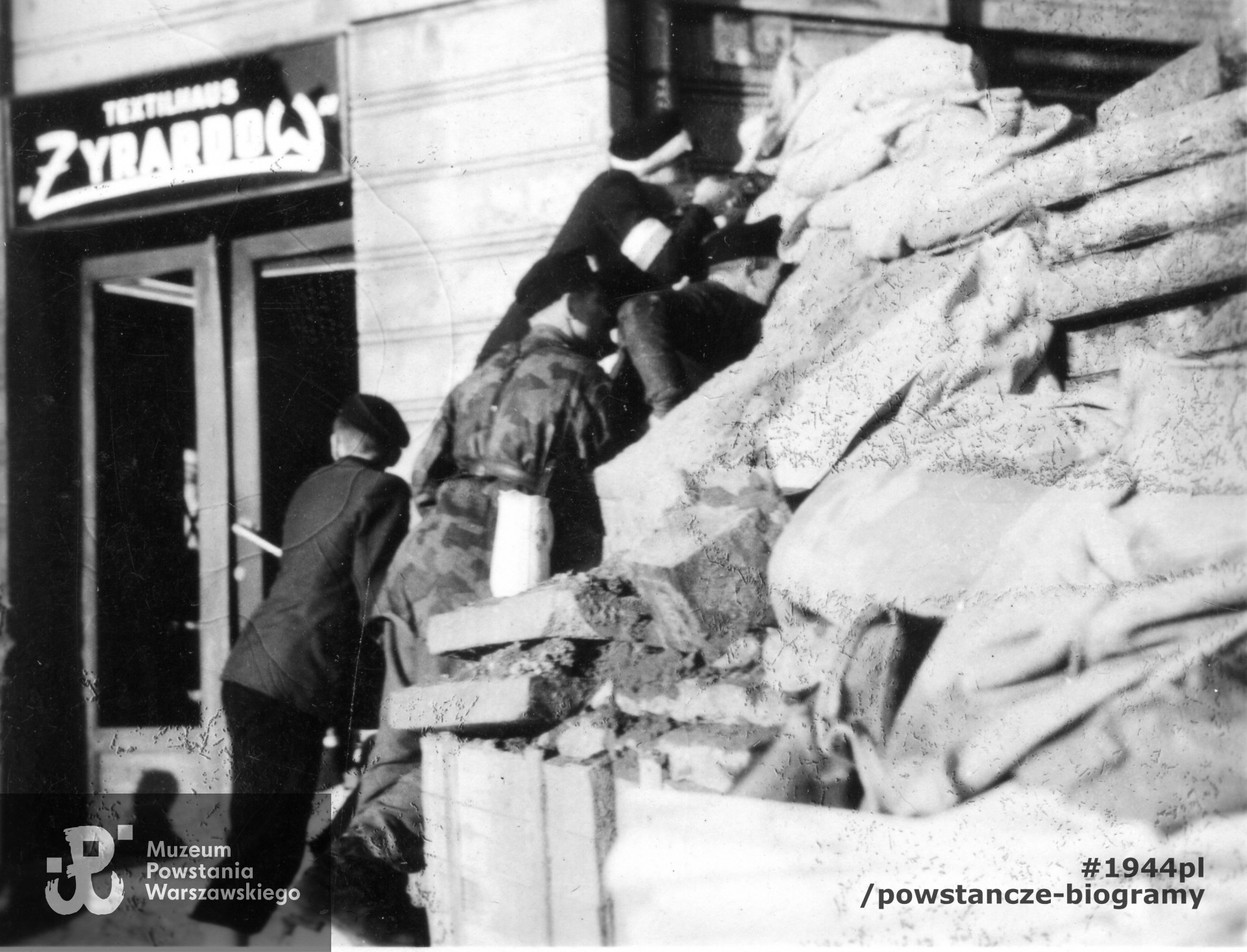 Fotografia z Powstania Warszawskiego. Starówka. Powstańcy przy barykadzie „Żyrardów”, zwanej też redutą „św. Trójcy” u wylotu ul. Daniłowiczowskiej na Bielańską. W tle widoczne wejście do sklepu firmowego fabryki tekstyliów „Żyrardów” przy ul. Bielańskiej 8.  Autorem zdjęcia jest Edward Tomiak. Fot. ze zbiorów Muzeum Powstania Warszawskiego, sygn. MPW-IP/4945/107