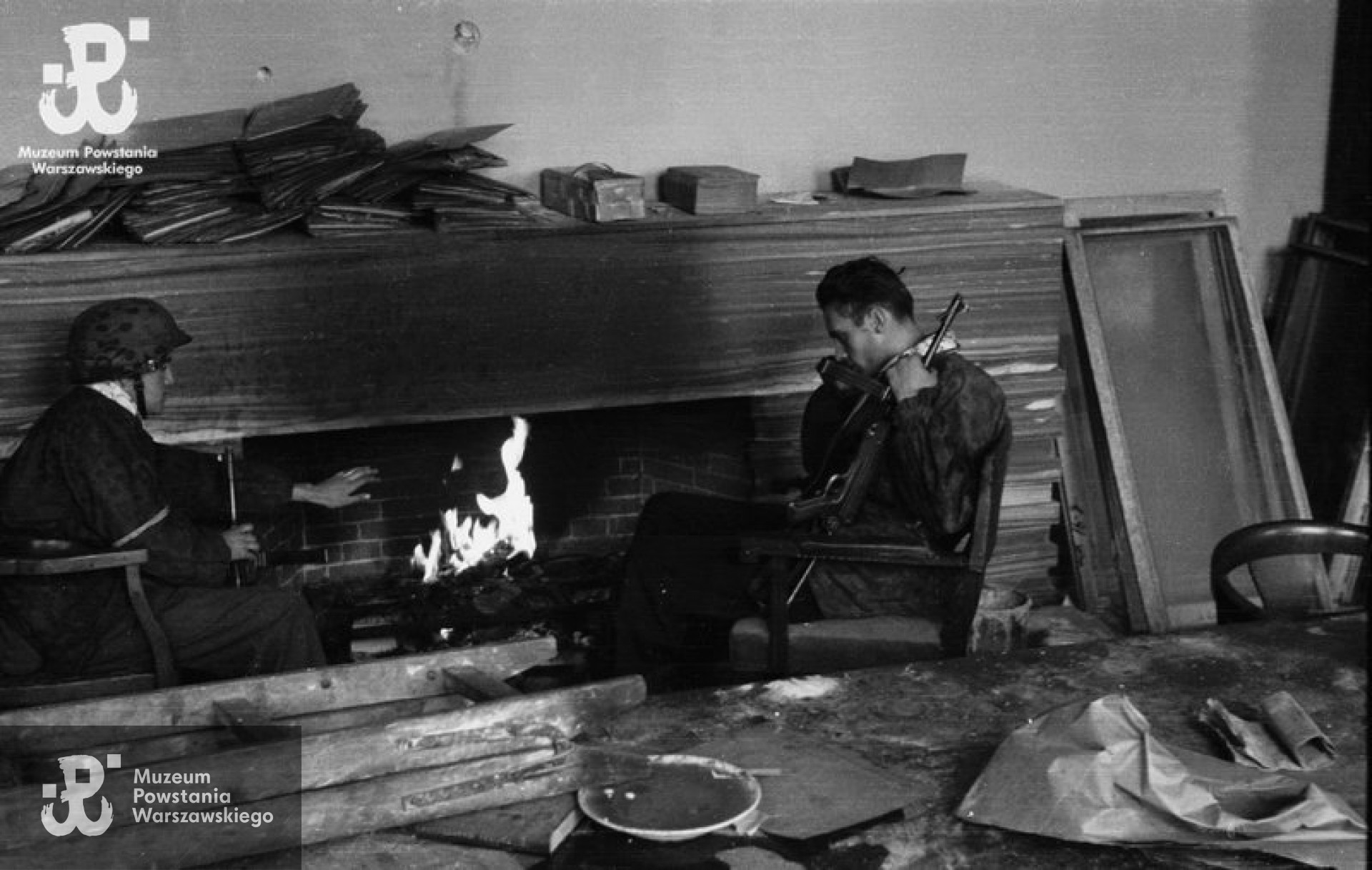 Fotografia z Powstania Warszawskiego. Śródmieście Północne, ulica Jasna, 1-2 października 1944.  Powstańcy z kompanii „Anna” Batalionu „Gustaw” przed kominkiem w gmachu Towarzystwa Ubezpieczeniowego „Polonia” przy ulicy Jasnej 19. Od lewej: kpr. Jerzy Sikorski „Sixton” , por. Wiesław Chrzanowski „Wiesław” z pistoletem maszynowym MP-40.  Fot. ze zbiorów Muzeum Powstania Warszawskiego, sygn. MPW-IN/3328