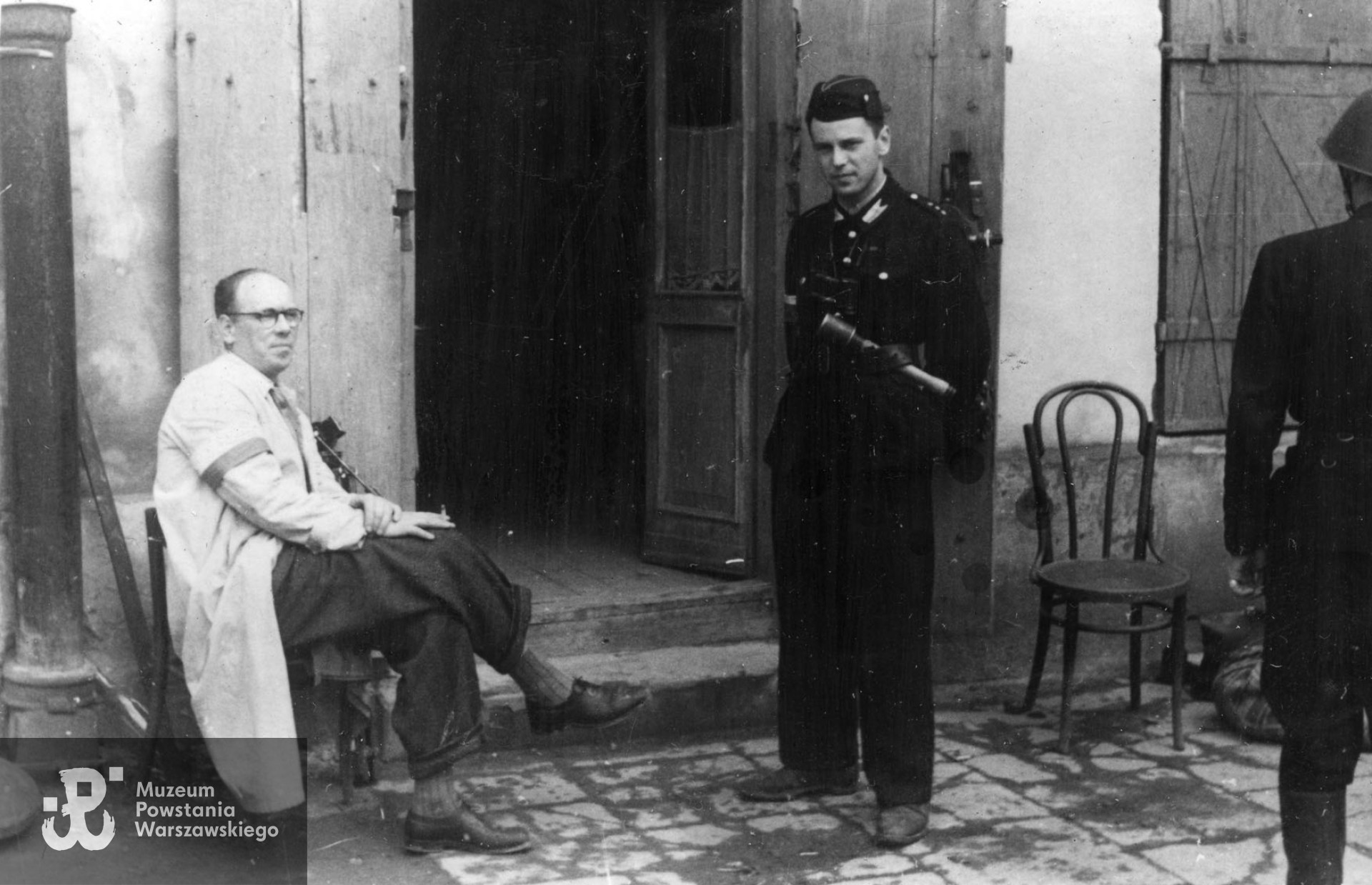 Fotografia z Powstania Warszawskiego autorstwa Bernarda Marwińskiego (PSW).  Śródmieście Północne, operator filmowy Henryk Vlassak siedzący na krześle przed wejściem do budynku. Na pierwszym planie tyłem Powstaniec z pistoletem. Fot. ze  zbiorów Muzeum Powstania Warszawskiego, sygn. MPW-IP/6832