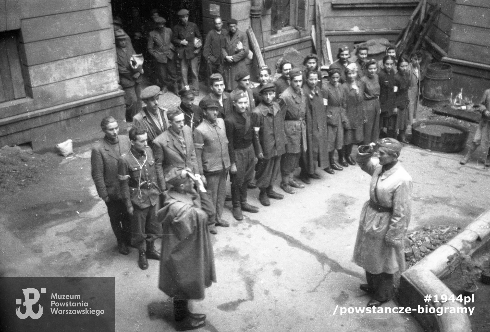 Fotografia z Powstania Warszawskiego. Powiśle. Zbiórka powstańców   ze zgrupowania "Krybar" w podwórzu kamienicy przy ulicy Tamka. W pierwszym rzędzie pierwszy z lewej Kazimierz Rupp "Dobiecki". Na rewersie odręczny opis autorstwa Kazimierza Ruppa: <i> 1944. Warszawa. Powstanie. Tamka. Ja w letnim mundurze ojca z wojny bolszewickiej 1920. </i> Ze zbiorów Muzeum Powstania Warszawskiego, sygn. P/4980-2. Ujęcie tożsame: MPW-IN/3691(P/5115/06 - zbiór Kazimierza Ruppa).
