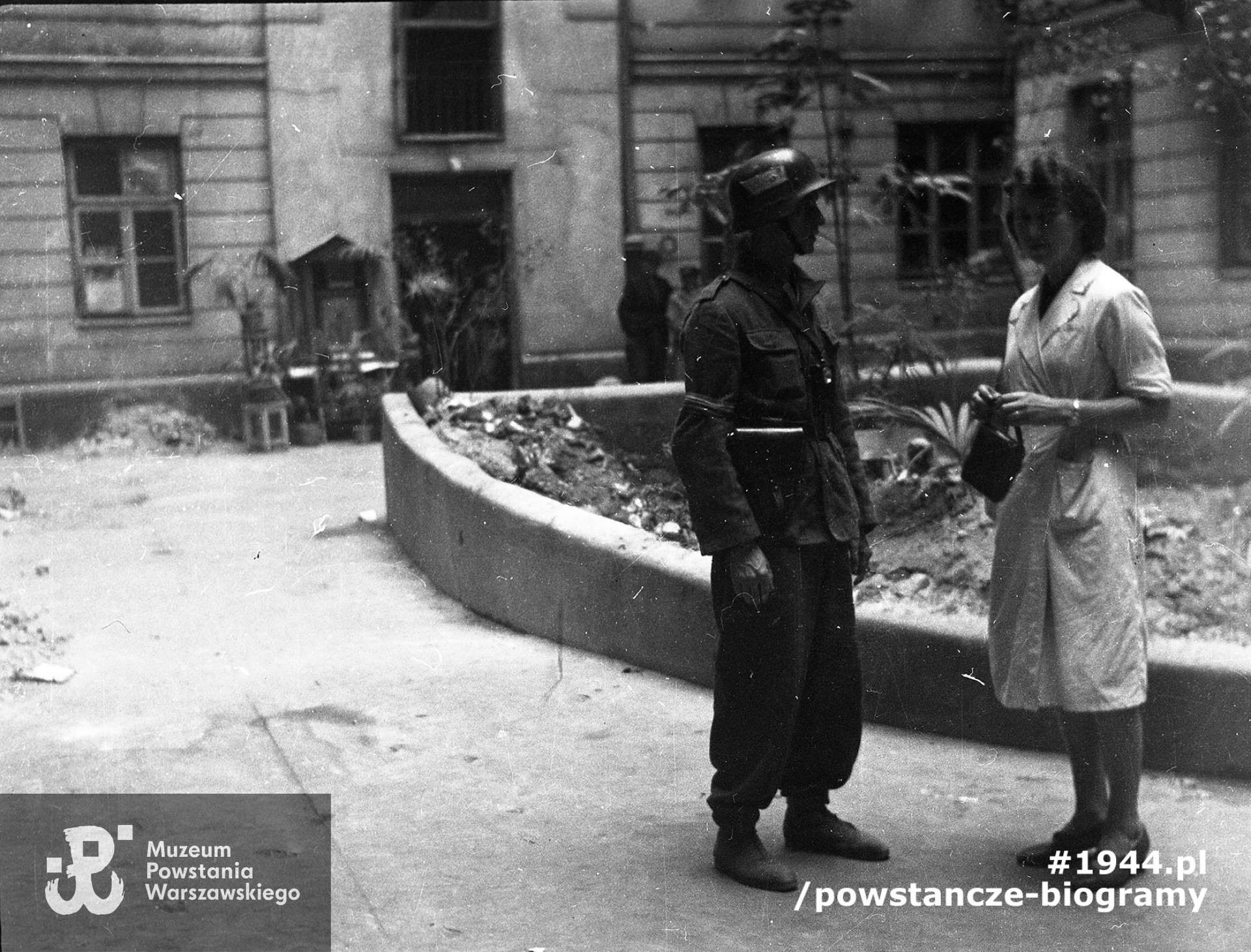 Fotografia z Powstania Warszawskiego autorstwa Zofii Wolskiej-Degórskiej. Sanitariuszka szpitala przy ul. Marszałkowskiej 40 Wanda Srzednicka "Aza" rozmawia z powstańcem na podwórku niezidentyfikowanej kamienicy. W drzwiach klatki schodowej stoją mężczyźni. Obok drzwi kapliczka. Fot. ze zbiorów Muzeum Powstania Warszawskiego, sygn. MPW-IN/10926