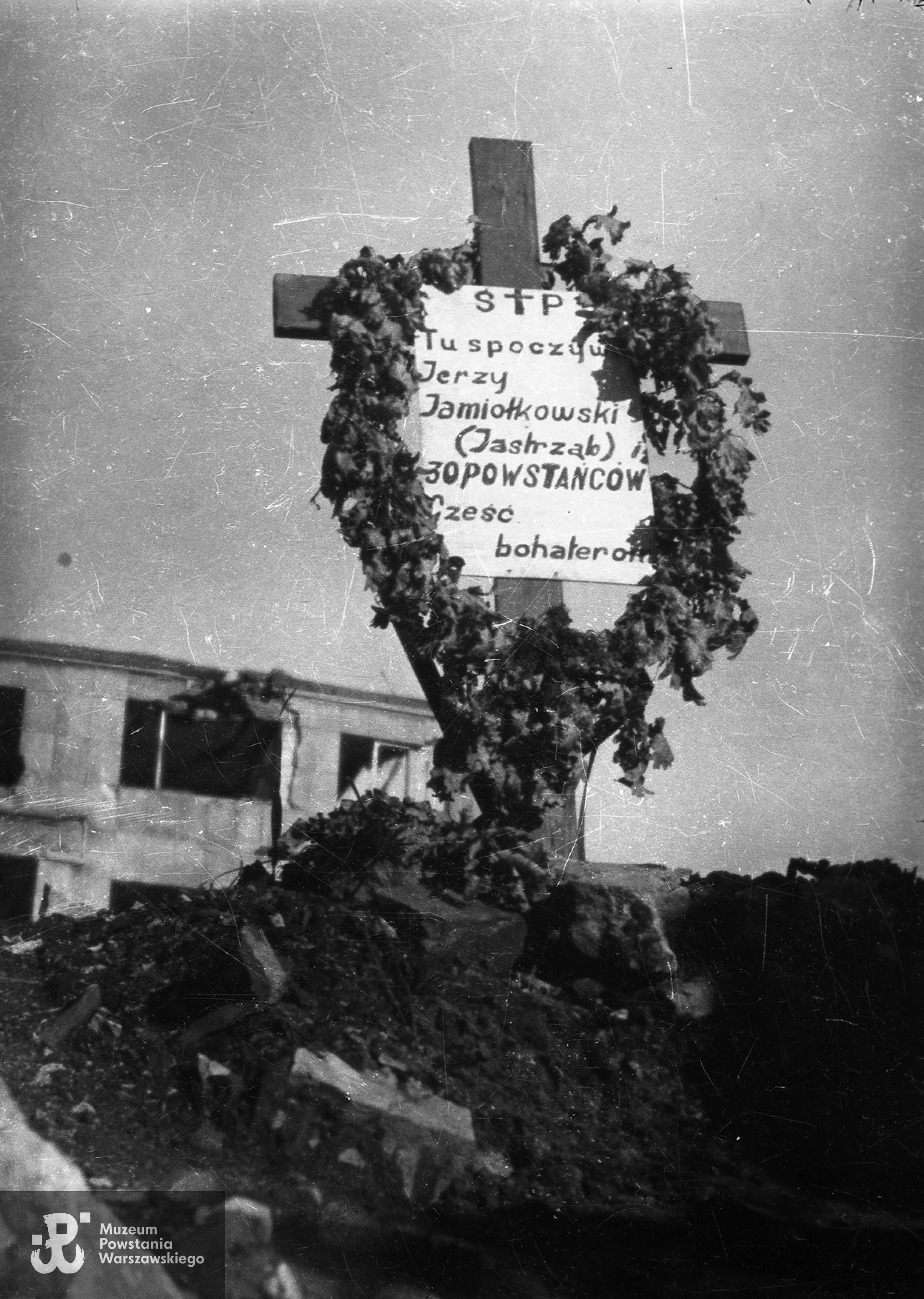 Fotografia z okresu powojennego. Warszawa. Grób kpr. Jerzego Stanisława Jamiołkowskiego "Jastrzębia" i powstańców poległych 2 września 1944 w gruzach zbombardowanego budynku Gimnazjum im. Królowej Jadwigi przy pl. Trzech Krzyży 1. Ujęcie w kierunku wschodnim. W tle po lewej widoczny fragment budynku górnych pięter  Riunione Adriatica di Sicurta przy pl. Trzech Krzyży 2. Na tabliczce inskrypcja: "Ś P Tu spoczywa Jerzy Jamiołkowski (Jastrząb) i 30 powstańców Cześć bohaterom". Fot. ze zbiorów Muzeum Powstania Warszawskiego, sygn. MPW-IN/9393