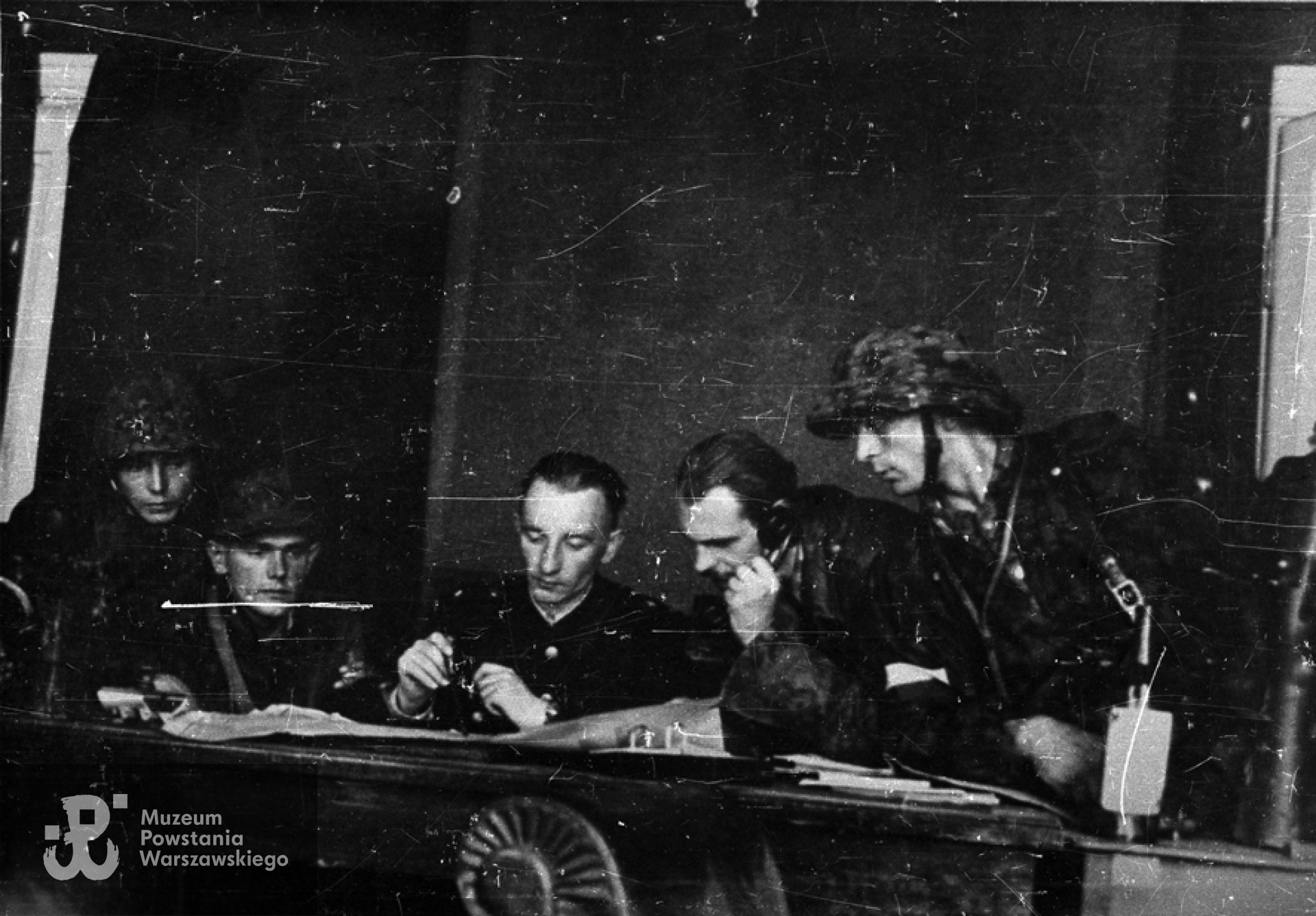 Fotografia z Powstania Warszawskiego. Stare Miasto. Odprawa poświęcona sytuacji na terenie zajętym przez 1 kompanię szturmową batalionu "Chrobry I". Od lewej: 1), pchor. Bogdan Czapliński "Wrzos", 2), "Czarny", 3) por. Eugeniusz Glass-Brudziński "Marek" - zastępca dowódcy 1. kompanii, 4) ppor. Jan Nestrypke"Boreysza" - dowódca 1. plutonu 1. kompanii i 5 N.N. Zdjęcie wykonane na kwaterze w Pasażu Simonsa, róg ul. Długiej i Nalewek. Autorem fotografii jest Zygmunt Sowiński "Ostoja". Ze zbiorów Muzeum Powstania Warszawskiego, sygn. MPW-IN/8702