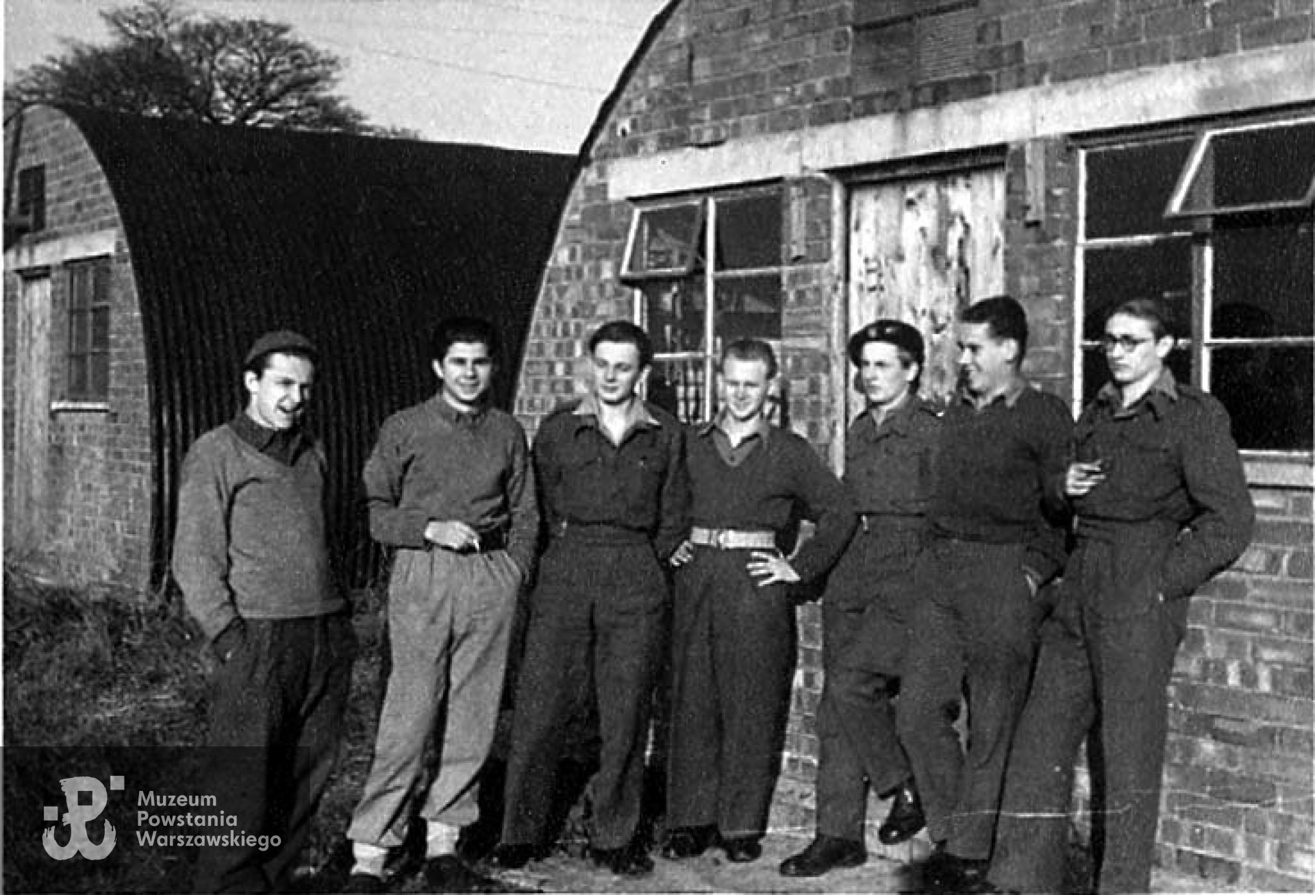 "Beczki śmiechu" (Nissen Huts) stanowiły pomieszczenia zarówno sal szkolnych, świetlic jak i sypialni, w których kwaterowało po dwunastu ludzi na barak. Krzysztof Głuchowski (w berecie) oparty o drzwi, z kolegami z baraku. Bodney Airfield Camp, jesień 1946 roku. Fot. z archiwum rodzinnego Krzysztofa Głuchowskiego.