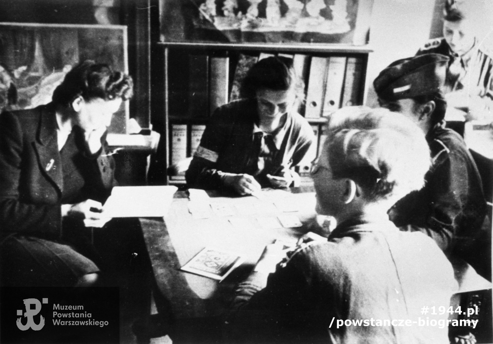 Fotografia z Powstania Warszawskiego autorstwa Joachima Joachimczyka. Kantor Zakładów Graficznych, chwila odpoczynku. w  Od lewej: Maria Zarembina, mieszkanka Chmielnej, łączniczki "Kowalska" (Teresa Włodzimierska), w furażerce "Boy" (Lucyna Piotrowska), tyłem "Kim" (Irena Glass). Przy oknie najprawdopodobniej Janina Wewiórska „Zawadzka” (?). Fot. ze zbiorów Muzeum Powstania warszawskiego, sygn. MPW-IH/2469