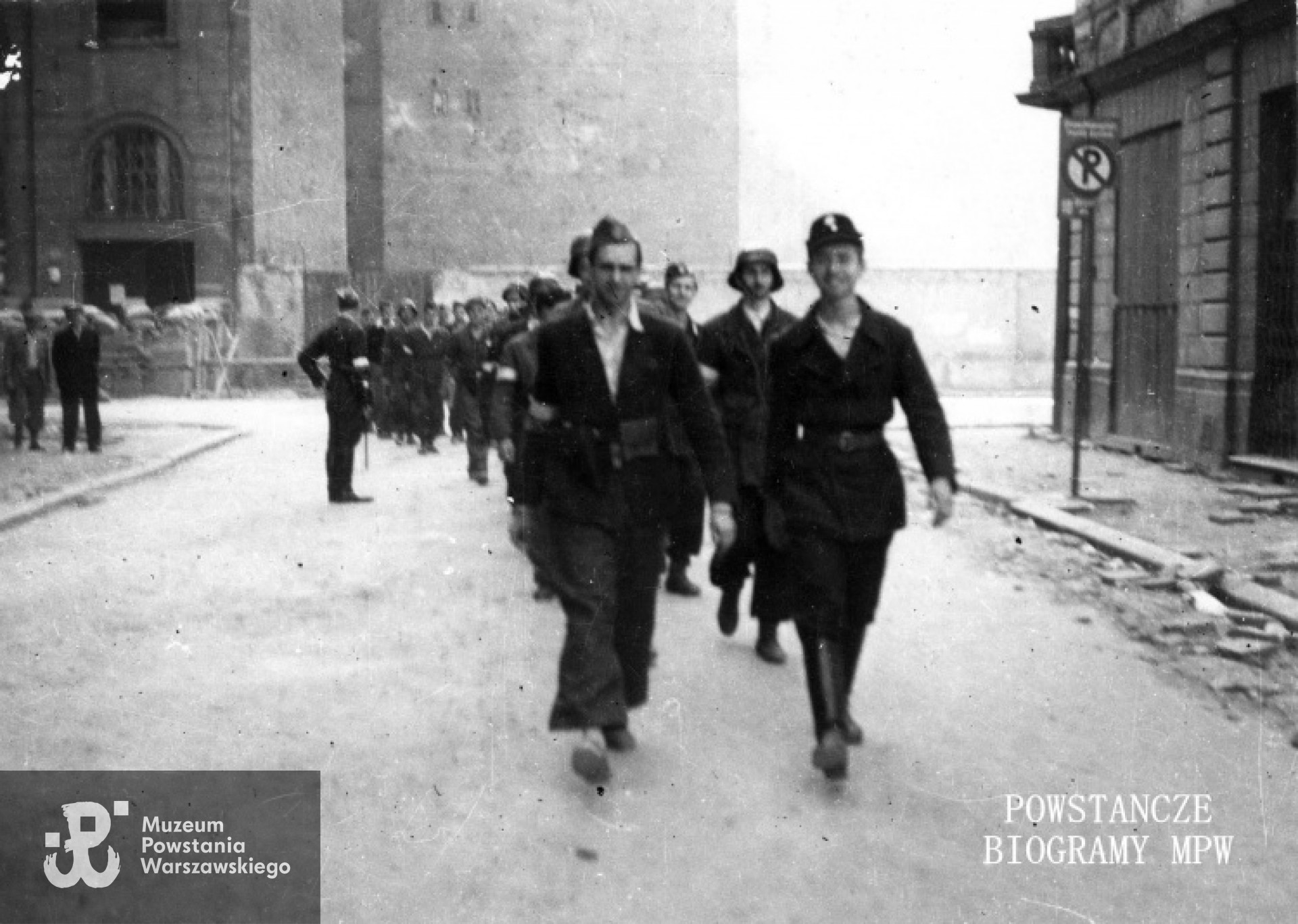 Fotografia z Powstania Warszawskiego. Wymarsz plutonu "Rygla" zgrupowania "Bartkiewicz" ul. Szkolną na akcję w Ogrodzie Saskim. W tle płd. pierzeja Świętokrzyskiej, po lewej widoczny fragment gmachu PKO Świętokrzyska 31/33 róg Jasna 9. Po prawej na pierwszym planie narożnik kamienicy Świętokrzyska 42 róg Szkolna 1/3. W pierwszej dwójce idzie kpr. pchor. Janusz Sawicki "Zabawa" (po prawej) z kpr. z cenz. Jerzym Jenike "Zwierzem" , w drugiej strz. Władysław Mydell "Waldemar" (po prawej) z  kpr. pchor. Andrzejem Glińskim "Leszczycem", w trzeciej z prawej kpr. pchor. Andrzej Strawiński "Chimera". Autor zdjęcia Irena Kummant-Skotnicka "Janina". MPW - sygn. MPW-IP/3844