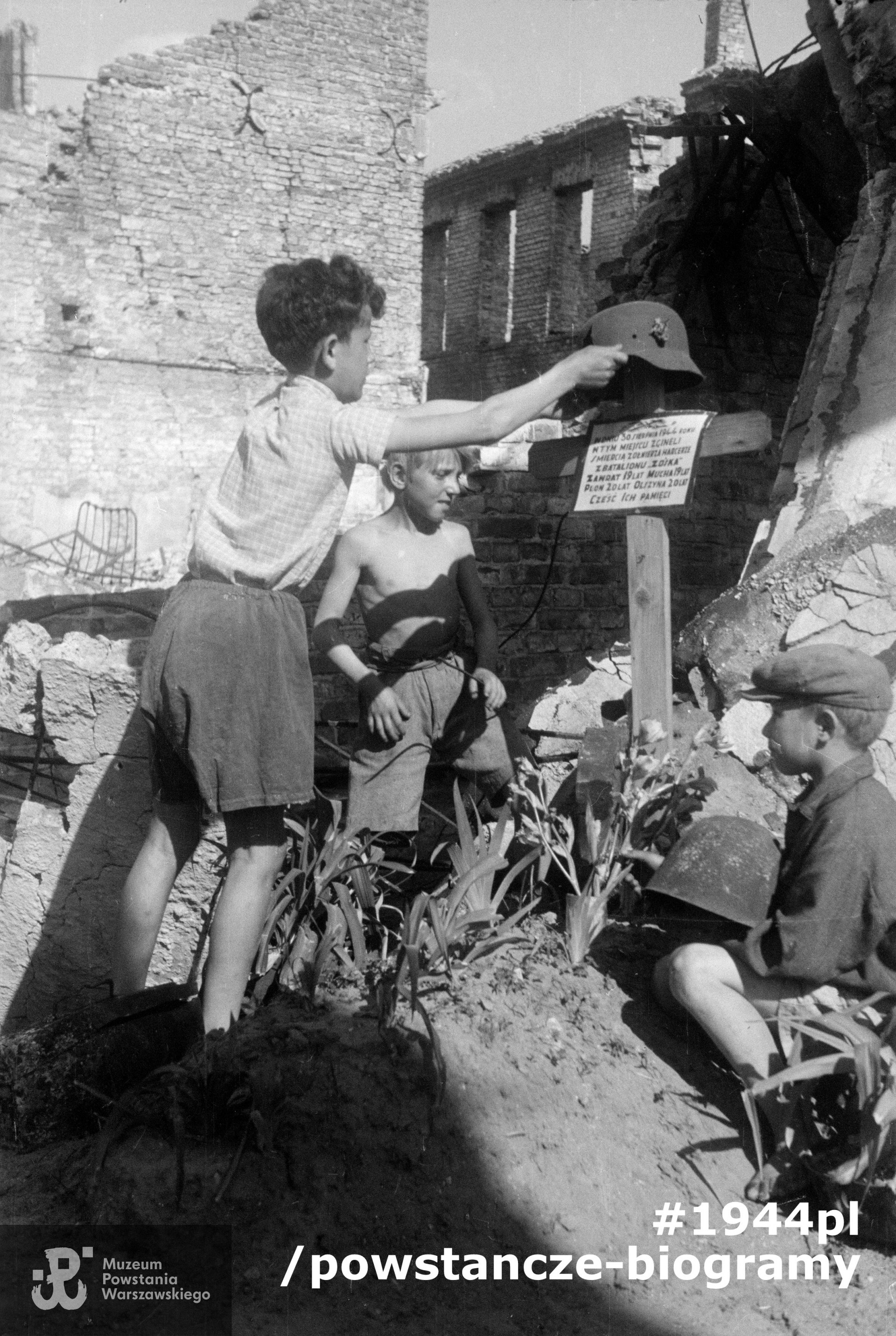Trzech chłopców przy grobie poległych harcerzy na terenie południowej hali zakładów Polskiego Fiata przy ul. Sapieżyńskiej 6. W tle najprawdopodobniej ruiny oficyn na posesji Zakroczymska 7 i Zakroczymska 5. Na grobie krzyż z założonym uszkodzonym hełmem (trzymanym przez chłopca), poniżej tablica z napisem: "W dniu 30 sierpnia 1944 roku w tym miejscu zginęli śmiercią żołnierza harcerze z batalionu "Zośka" Zawrat [Tadeusz Muller] 19 lat Mucha [Stefan Wiśniewski] 19 lat Plon [Zbigniew Wojciechowski] 20 lat Olszyna [Stanisław Findeisen] 20 lat Cześć ich pamięci".  Fot. ze zbiorów Muzeum Powstania Warszawskiego, sygn.  MPW-IN/12472