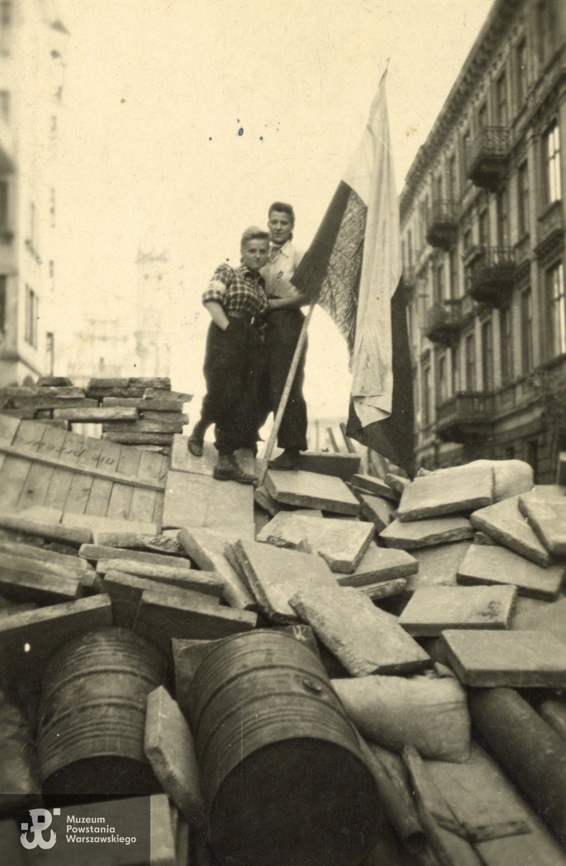 Fotografia wykonana w trakcie Powstania Warszawskiego w Śródmieściu Południowym na barykadzie na ul. Mokotowskiej, widok w kierunku Placu Zbawiciela. W tle widoczna po lewej wieża kościoła św. Zbawiciela. Na zdjęciu dwie sanitariuszki - Zofia Zych i Jadwiga Kostrzanowska. Na rewersie dedykacja dla kolegi: "Ciężko przystojnemu, diabelnie miłemu, Zosia. Darmstadt, 9 X 1945". Na dole dopisek: Zofia Zych, Jadwiga Kostrzanowska, zginęła w Powstaniu".  Adresatem dedykacji był najprawdopodobniej Jerzy Działak ps. "Hińcza". Fot. pochodząca ze spuścizny po Jerzym Działaku, obecnie w zbiorach Muzeum Powstania Warszawskiego, dar p. Anny Działak-Szubińskiej.