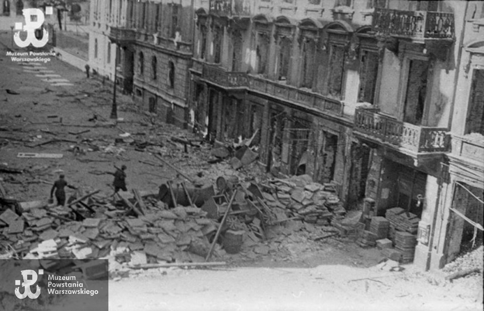 Fotografia z Powstania Warszawskiego. Okolice "Reduty Kuczyńskich" - Śródmieście Południowe. Barykada na ul. Brackiej róg ul. Nowogrodzkiej (pomiędzy Bracka 9 i 12), w głębi skrzyżowanie Brackiej i Al. Jerozolimskich (widoczny murek z metalowym parkanem, ogradzał ogródek przylegający do BGK - pozycje niemieckie) Po drugiej stronie Alej Jerozolimskich - fragment ruin budynku (Bracka 16), w którym na parterze znajdowała się przed wojną znana restauracja "Cristal". W bramie kamienicy Bracka 12 powstańcy wybudowali z płyt chodnikowych "bunkier". "Reduta Kuczyńskich" była zlokalizowana w narożnym domu Bracka 13, w którym przed wojną mieściła się kawiarnia Braci Kuczyńskich. Fot. ze zbiorów Muzeum Powstania Warszawskiego.
