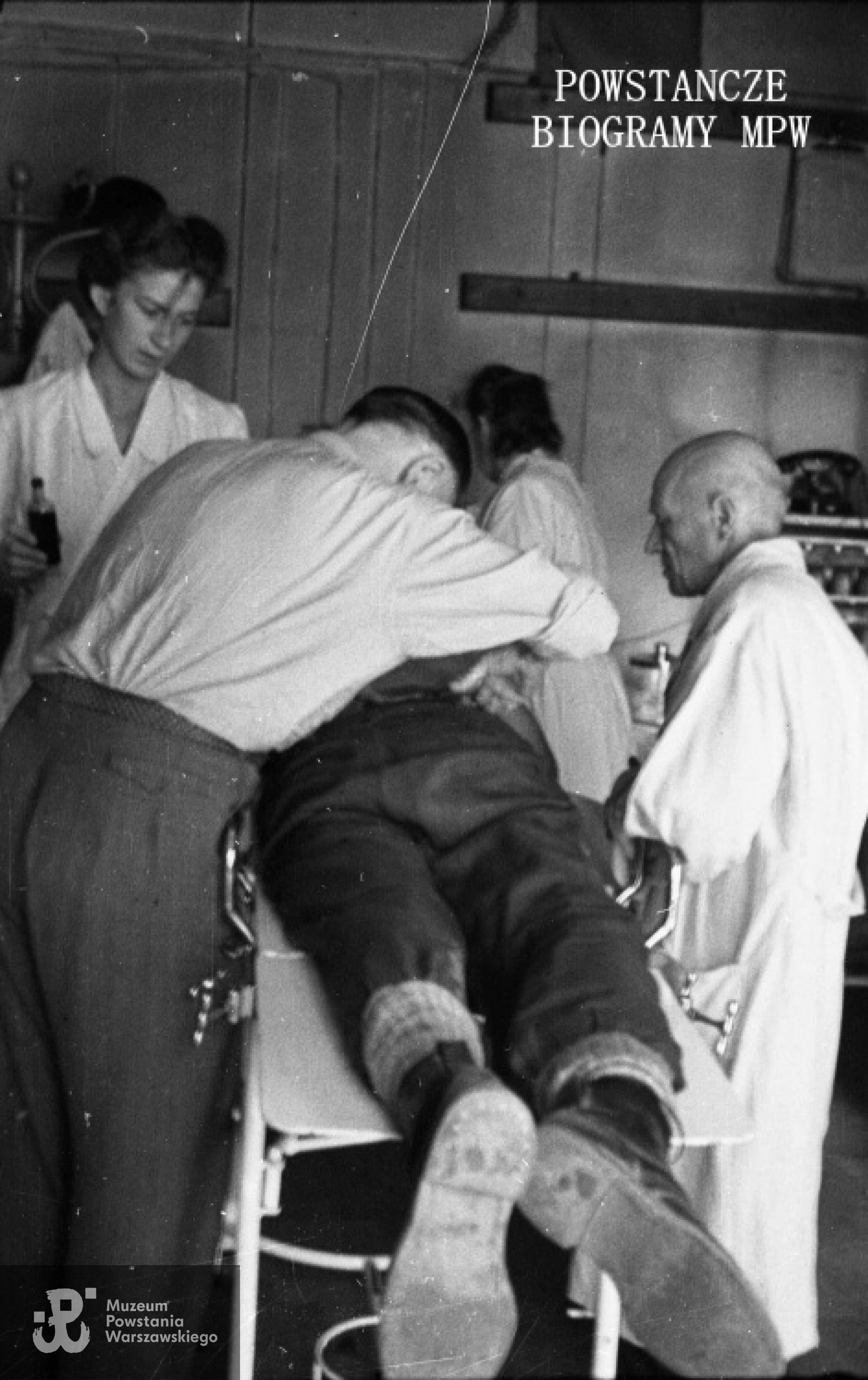 Fotografia z Powstania Warszawskiego. Ranny powstaniec opatrywany w Szpitalu Polowym nr 1 na ul. Wspólnej 27. Po prawej widoczny w białym fartuchu ppłk.dr Tadeusz Bętkowski "Tata". Fot. ze zbiorów Muzeum Powstania Warszawskiego, sygn. MPW-IN/1537