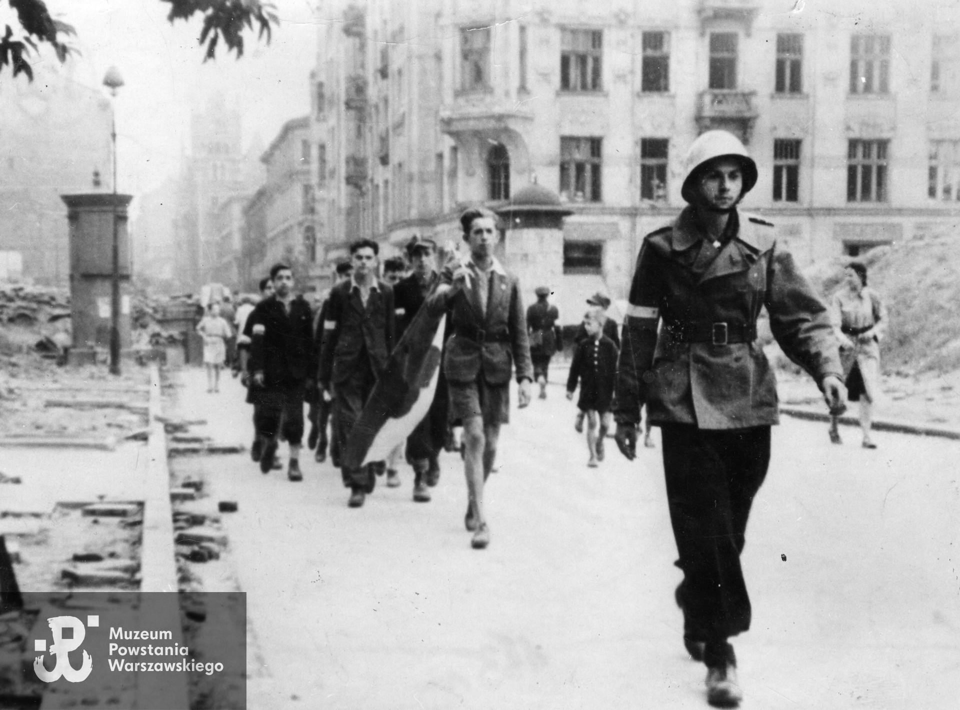 Fotografia z Powstania Warszawskiego. Śródmieście Północne Ul. Sienna. Oddział powstańczy z flagą maszerujący ulicą Sienną. Ujęcie spod kamienicy nr 14 (poza kadrem) w kierunku wschodnim. W tle po prawej kamienica przy ulicy Siennej 5/7 róg Wielkiej 14. Fot. ze zbiorów Muzeum Powstania Warszawskiego, sygn. MPW-IK/1978. Według identyfikacji p. Zbigniewa Kruszewskiego z flagą kroczy Jerzy Duczkowski „Juras” (1927-1954), a za nim widoczny jest po lewej Zbigniew Kruszewski „Jowisz".  Odbitka w zbiorach MPW nie jest opisana i osoby na zdjęciu nie są rozpoznane.