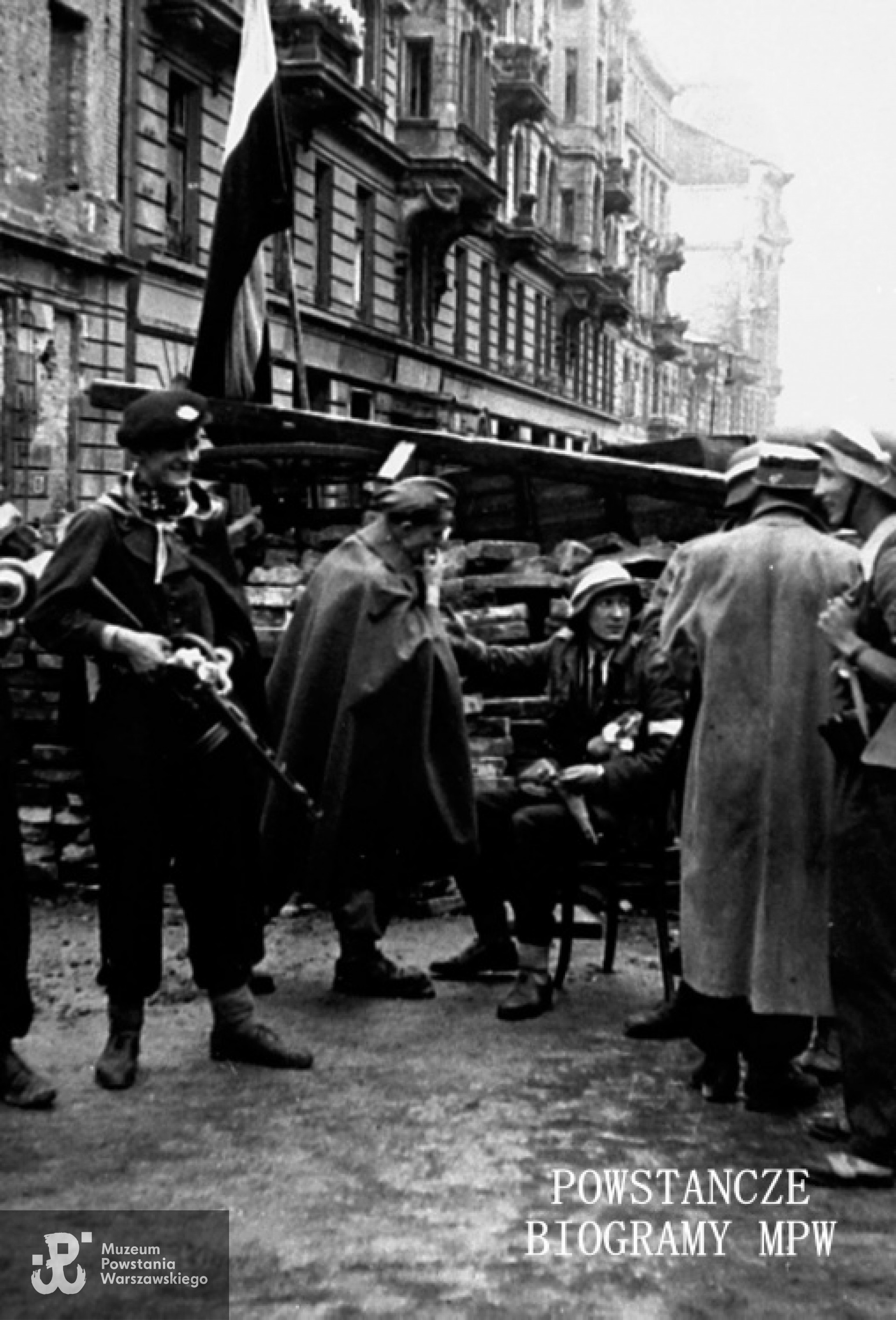 Fotografia z Powstania Warszawskiego. Śródmieście Północne. Obsada barykady przegradzającej ul. Bracką na wysokości kamienicy nr 20. Ujęcie w kierunku południowo-wschodnim. Powstańcy z 3 kompanii "Szare Szeregi" batalionu "Kiliński"; w pelerynie - Władysław Aniołek "Kot", na krześle siedzi - Maciej Lubicz "Bohdan", który poległ na tej barykadzie 4 sierpnia. Fot. ze zbiorów Muzeum Powstania Warszawskiego, sygn. MPW-IH/4208