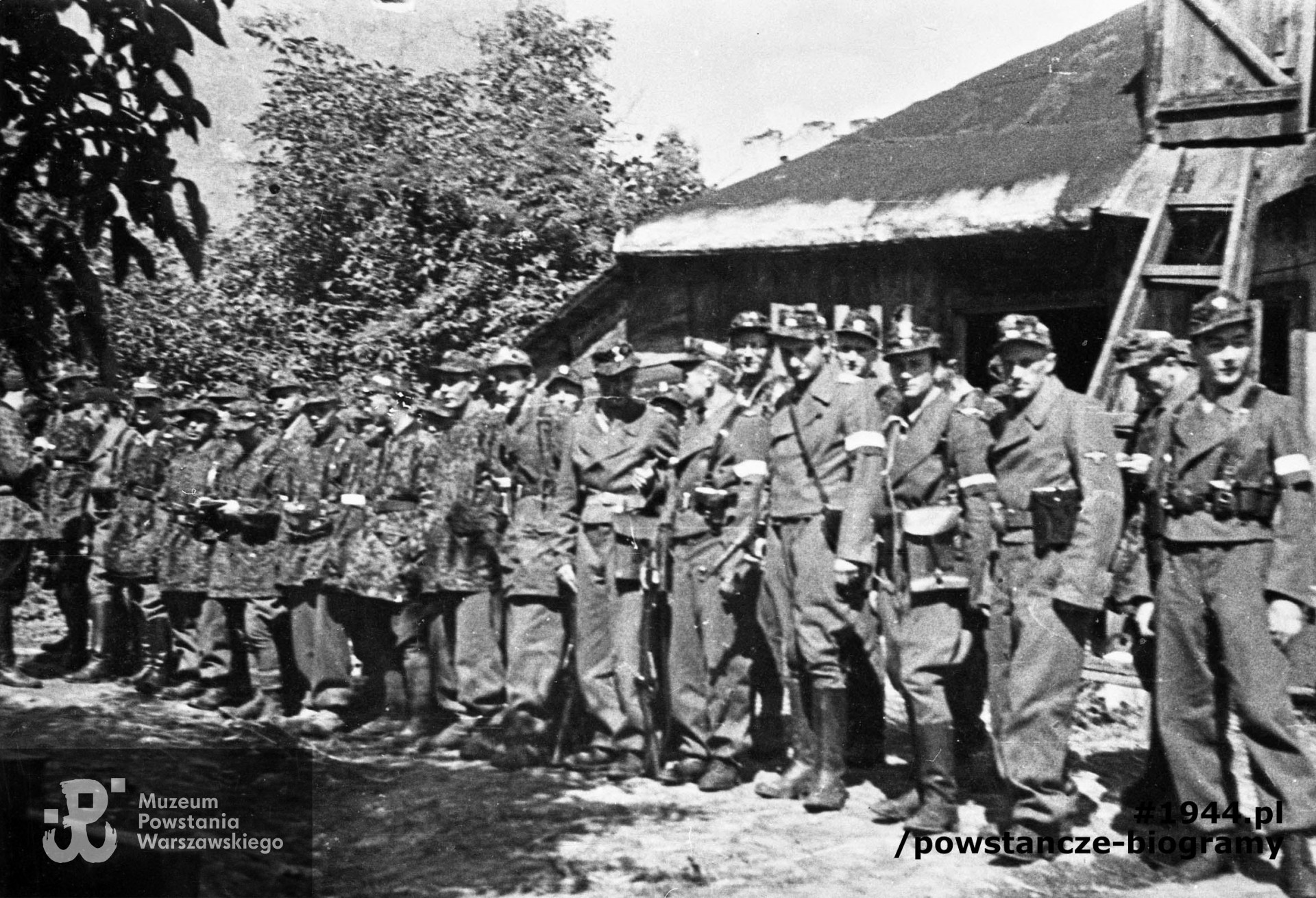 Fotografia z Powstania Warszawskiego. Wola. Żołnierze Kolegium "A" Kedywu OW AK w ogrodzie RGO - ul. Okopowa 41. Stoją od lewej: Aleksander Kabański "Brzozowski" (bokiem), trzeci - Jan Bagiński "Socha" (w berecie), czwarty - Zygmunt Siennicki "Bor" , ósmy - Józef Tomaszewski "Klemens". Dziewiąty od prawej - Jerzy Krzymowski "Jodła" (twarz w cieniu), czwarty od prawej- Jan Kulczycki "Argos" (w długich butach). Fot. ze zbiorów Muzeum Powstania Warszawskiego, sygn. MPW-IN/3643