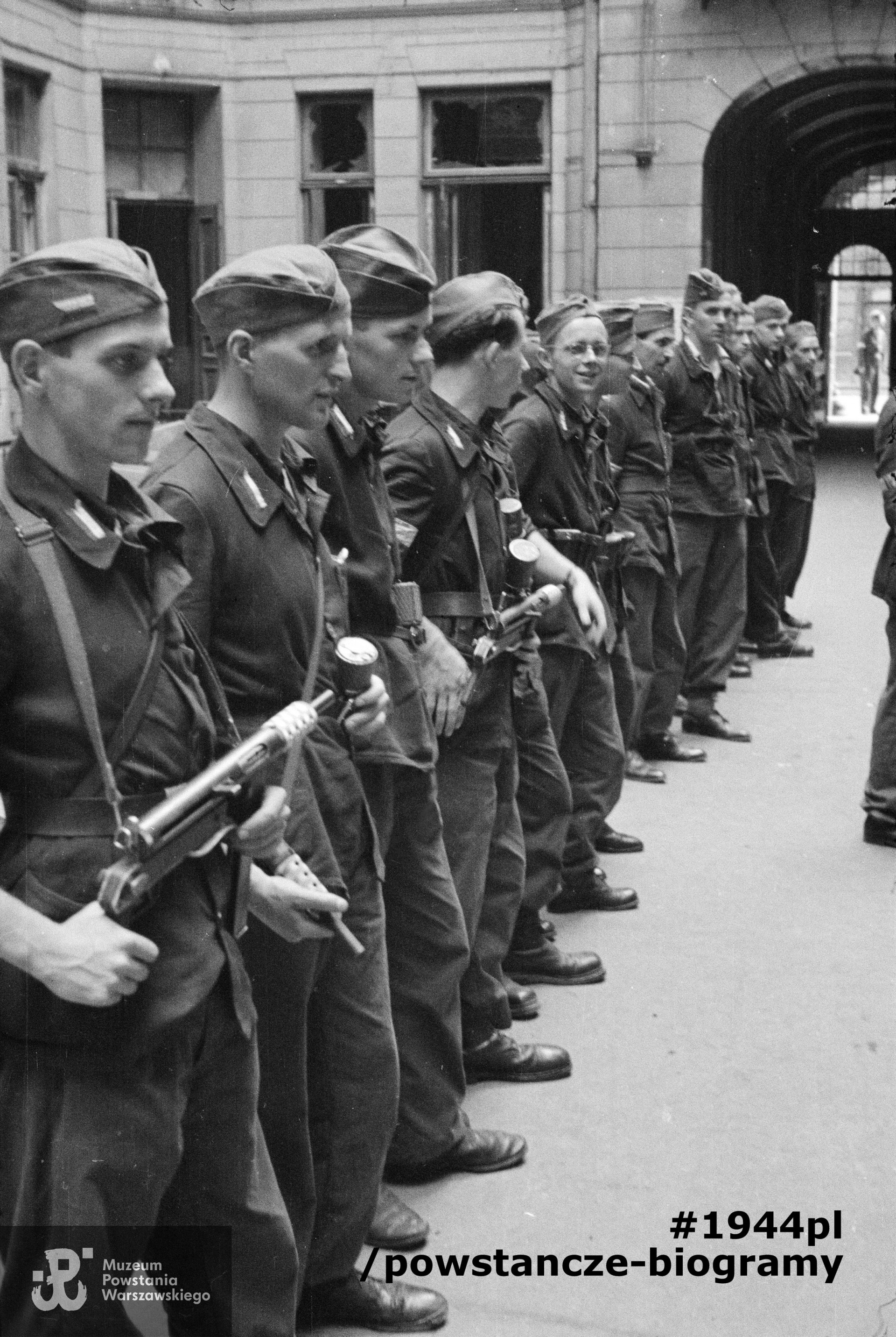 Fotografia z Powstania Warszawskiego. Śródmieście Północne. Apel III plutonu Oddziału Osłonowego Wojskowych Zakładów Wydawniczych (WZW) przy Oddziale VI Sztabu Komendy Głównej AK na podwórzu kamienicy Lewenfisza przy pl. Napoleona 1, róg ul. Boduena 2, róg ul. Sienkiewicza 1. Ujęcie w kierunku południowym. W tle widoczna brama wychodząca na ulicę Boduena. Stoją od lewej: ppor. Józef Kulesza "Mały Dżo", ppor. Władysław Wojtkiewicz "Ohm", st. strz. Jerzy Słubicki "Jarema", kpr. pchor. Tadeusz Bruszewski "Sztubak", kpr. Roman Cichalewski "Romek”, „Okularnik”, (NN), (NN), (NN), (NN), strz. Włodzimierz Leo "Bawół" i kpr. pchor. Ryszard Wrzosek "Dick". Autorem zdjęcia jest Eugeniusz Lokajski "Brok". Fot. ze zbiorów Muzeum Powstania Warszawskiego, sygn. MPW-IN/715