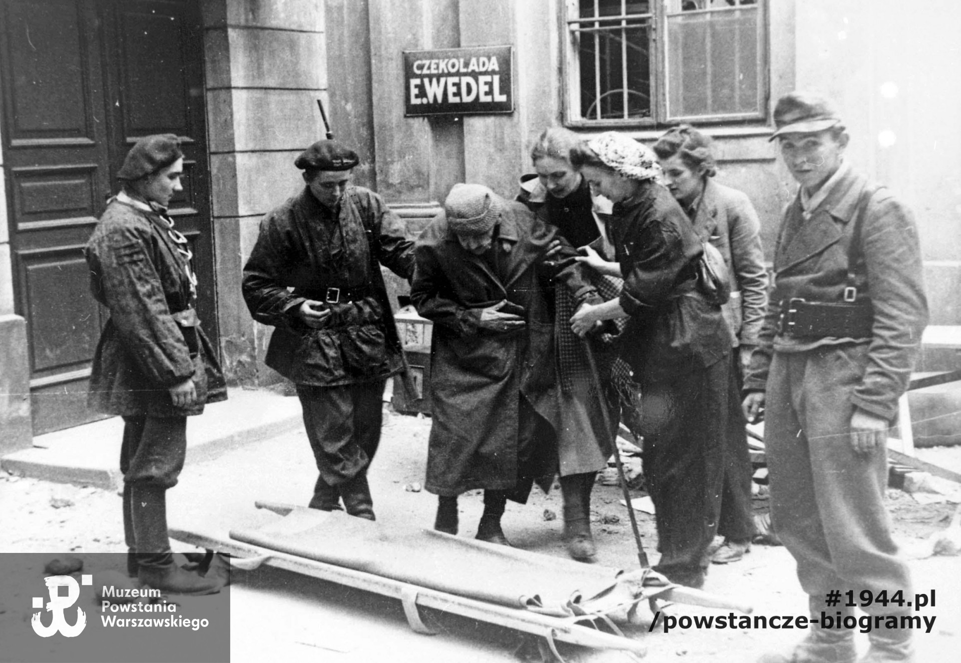 Fotografia wykonana podczas Powstania Warszawskiego. Śródmieście Północne. Patrol sanitarny z III Zgrupowania "Konrad" przed sklepem "Wedla", przy ul. Szpitalnej 8, podczas ewakuacji pisarki Marii Rodziewiczówny. Autorem zdjęcia jest Antoni Bohdziewicz. Fot. ze zbiorów Muzeum Powstania Warszawskiego, sygn. MPW-IP/3200