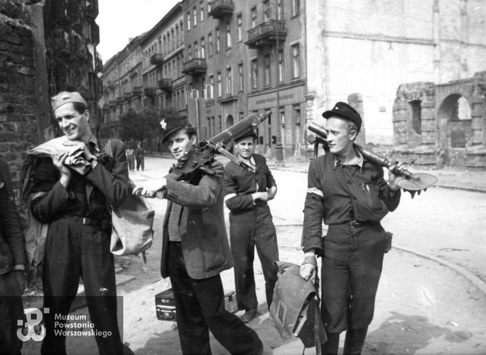 Zdobywcy Komendy Policji obładowani bronią na ul. Czackiego przy Sienkiewicza. Od prawej: rozebrana w czasie okupacji zniszczona kamienica Czackiego nr 2 (róg Świętokrzyska), dalej budynki nr: 4, 6, 8 i 10. Po prawej stoi strz. "Zimny" - Ryszard Lipski z trójnogiem do MG-08/15 Maxim.